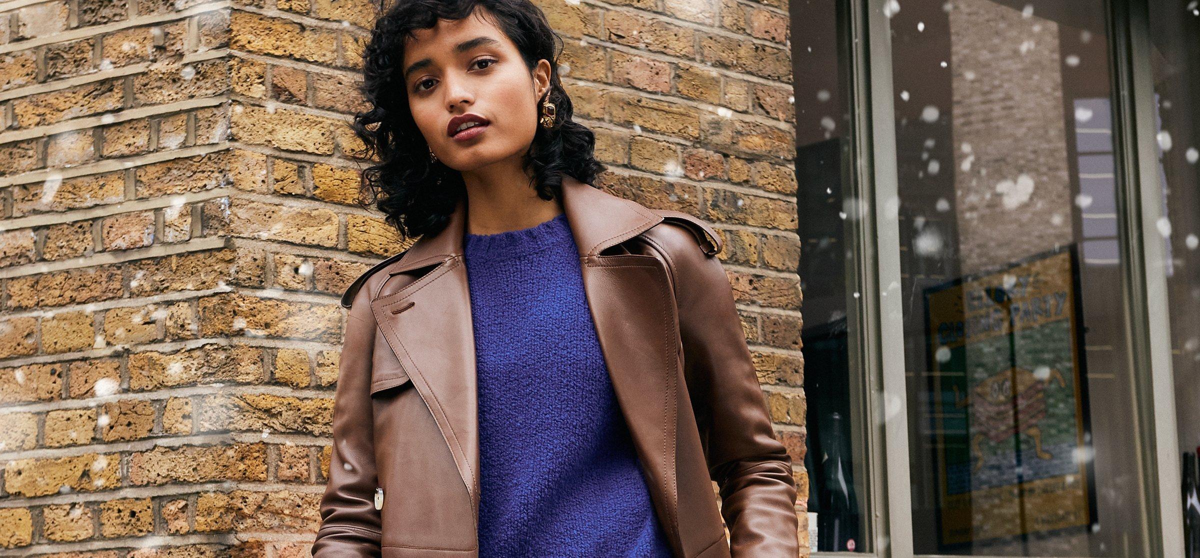 Image of a lady standing in front of a brick wall with snow falling down wearing a brown leather jacket and blue jumper