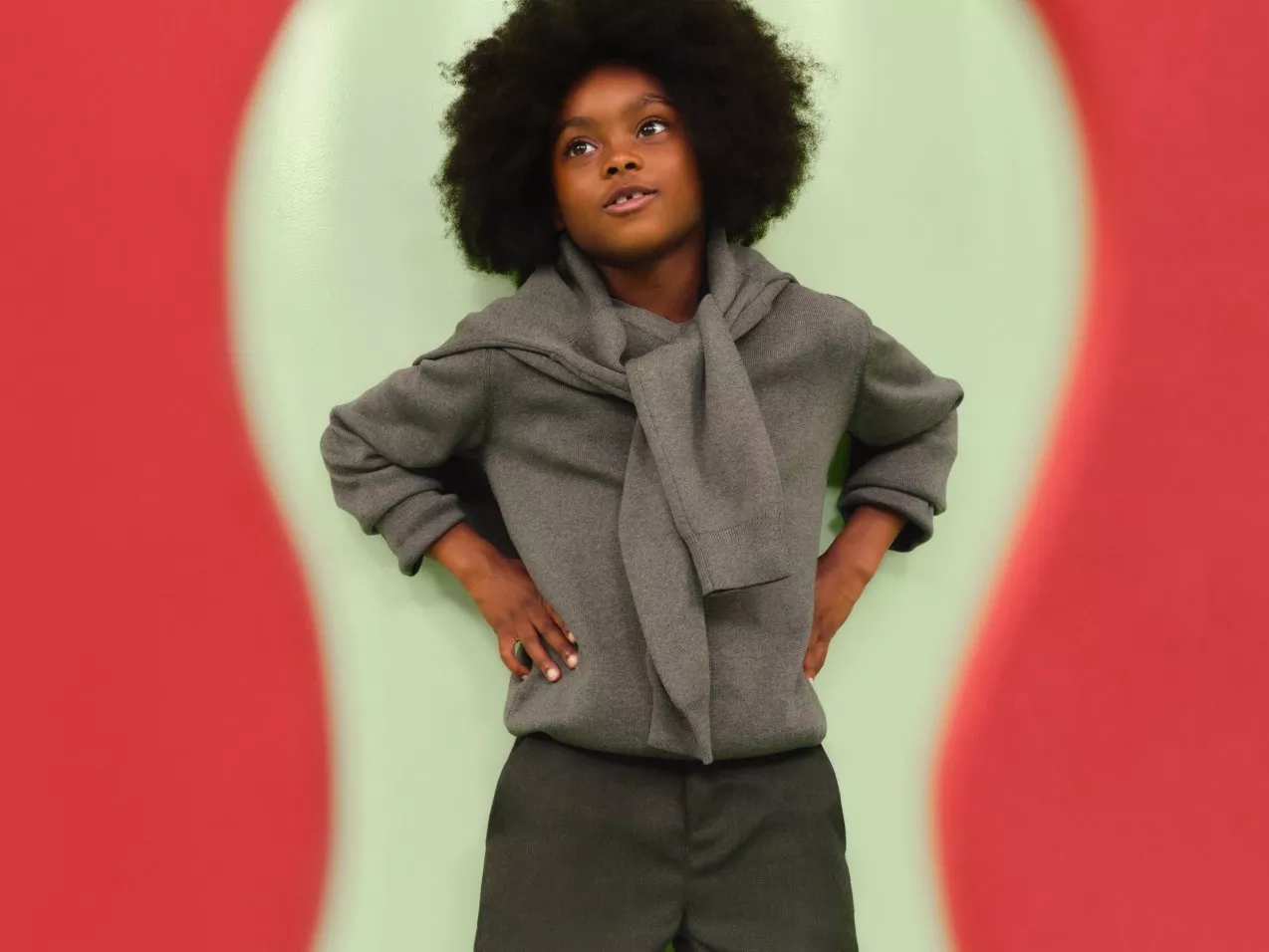 Stylish child in grey jumper and scarf, hands on hips, posing against bold red and mint studio backdrop