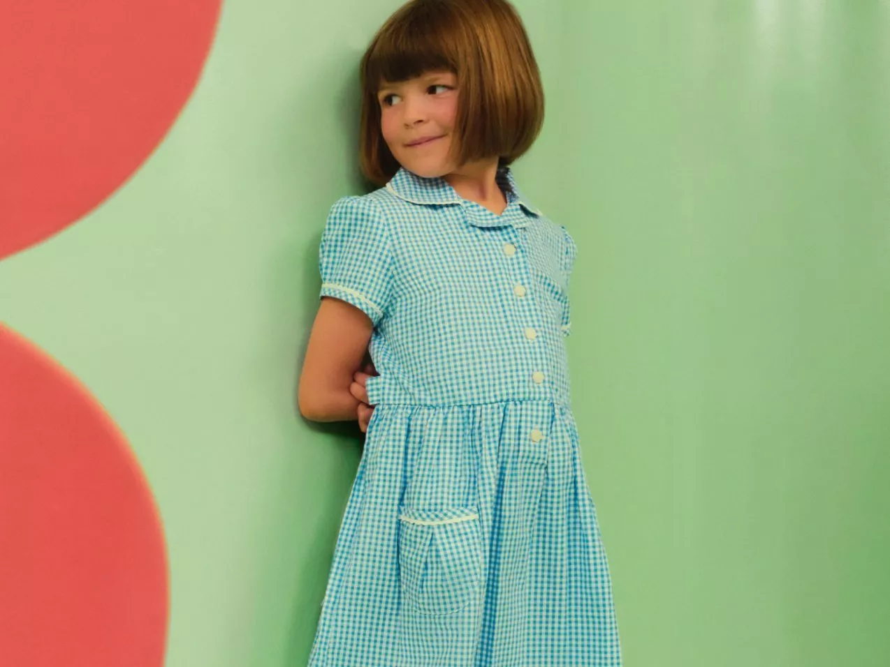 Smiling young girl in blue gingham school dress standing against green wall with pink circles, kids fashion portrait