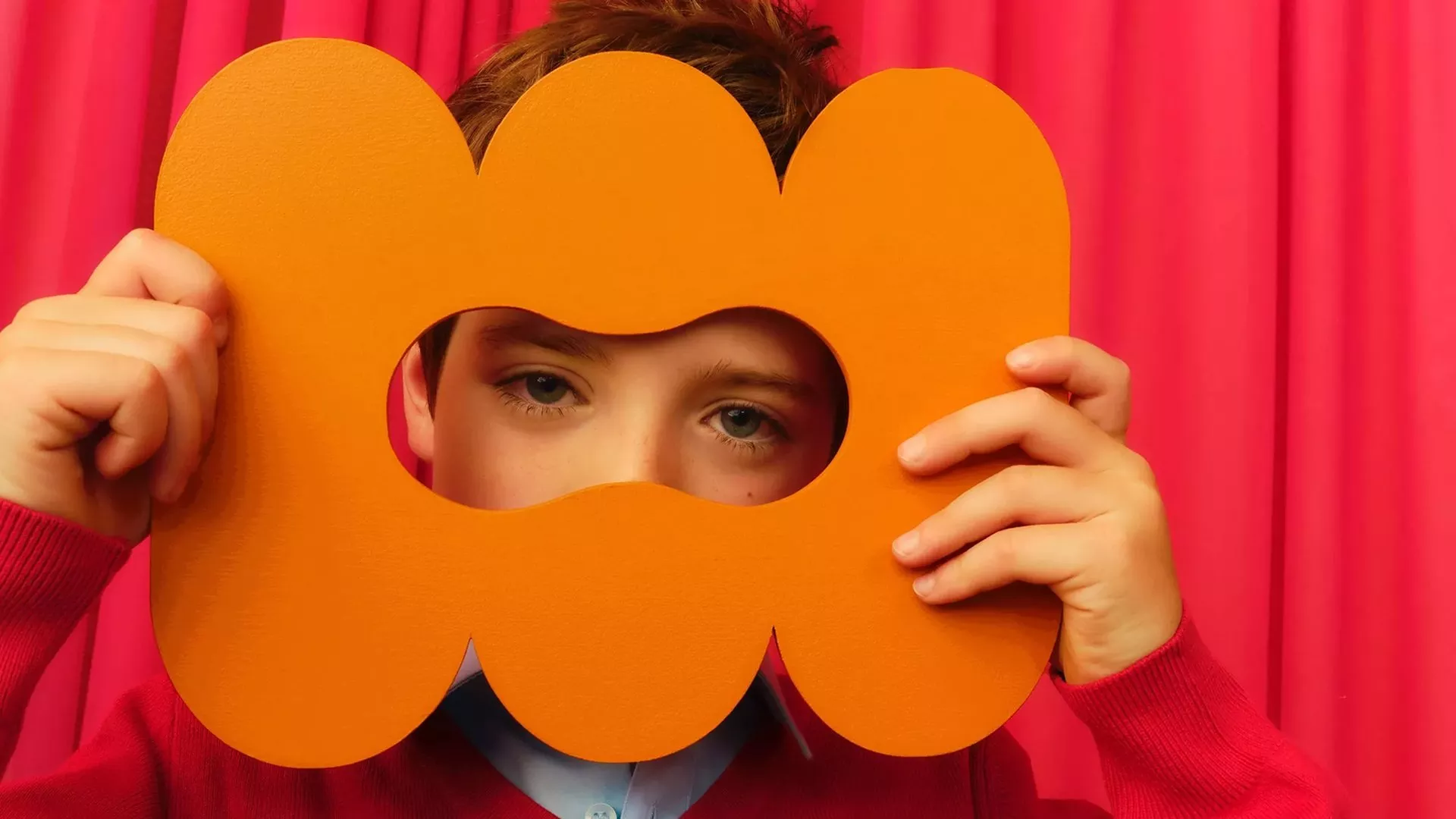 Boy holding orange flower-shaped photo booth prop frame over face against bright pink curtain backdrop