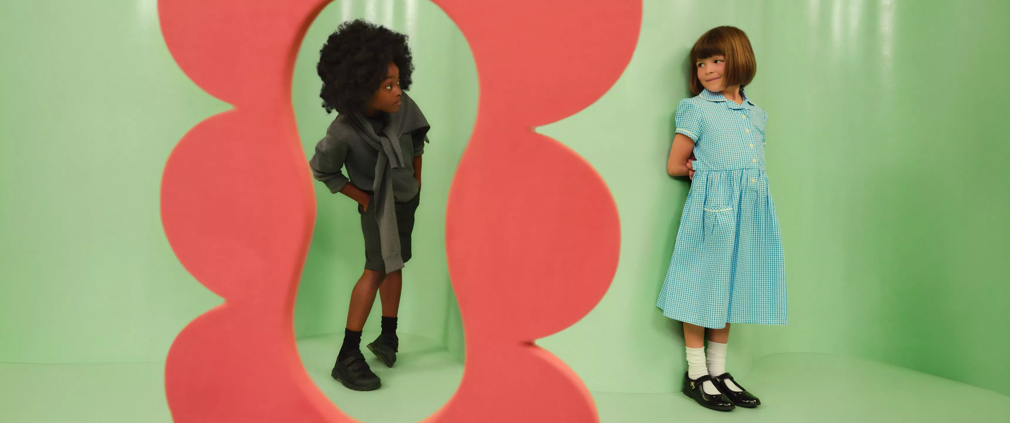 Two children in stylish outfits pose by a pink cut-out on a pastel green studio background, playful fashion shoot
