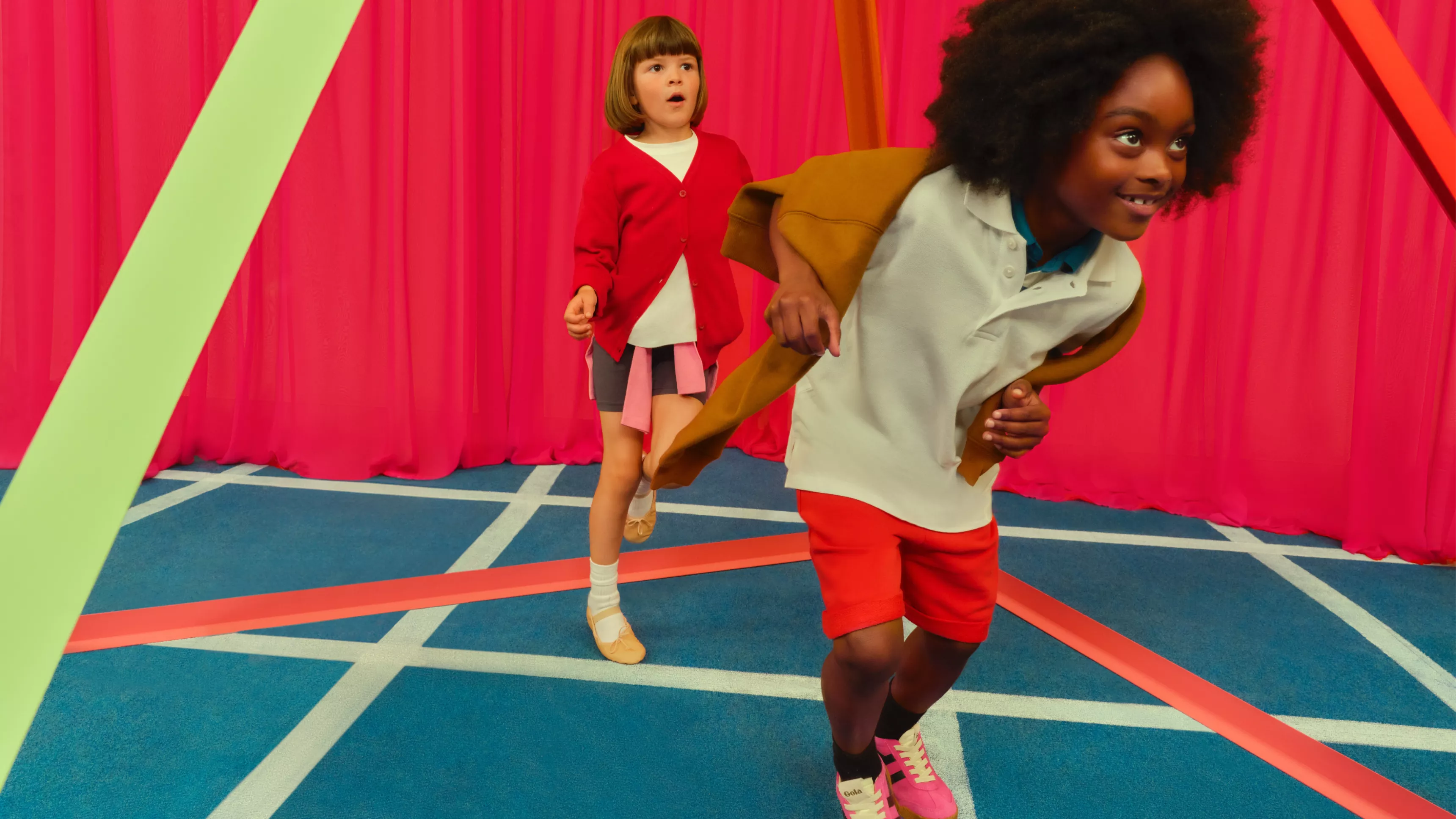 Two children in casual sportswear, running indoors on a colourful set with pink curtains and geometric lines on the floor
