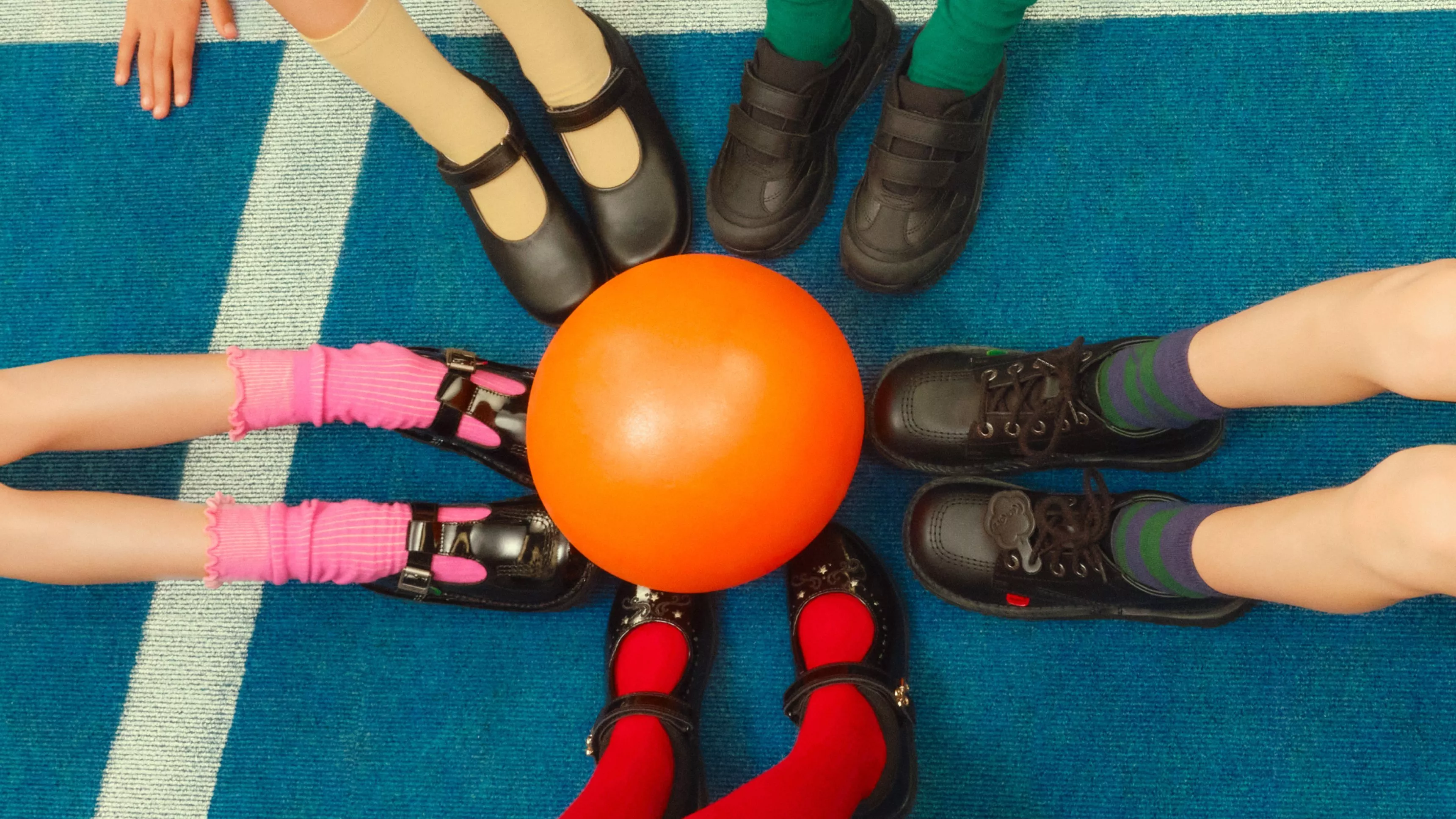 Children’s feet in school shoes around an orange ball on blue carpet, circle game in classroom or playground