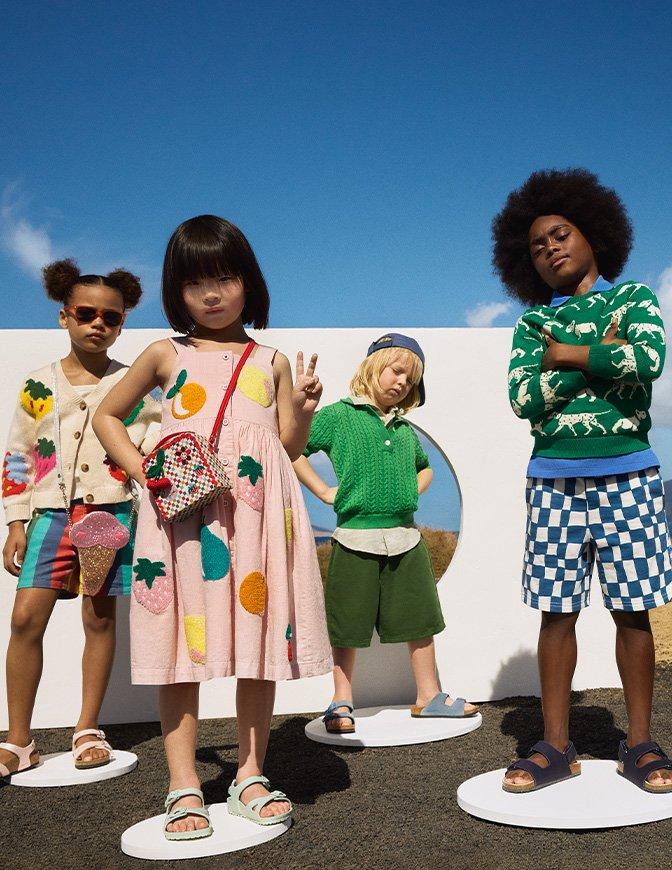 Four stylishly dressed children pose confidently on circular platforms outdoors against a blue sky, showcasing colorful and playful summer outfits