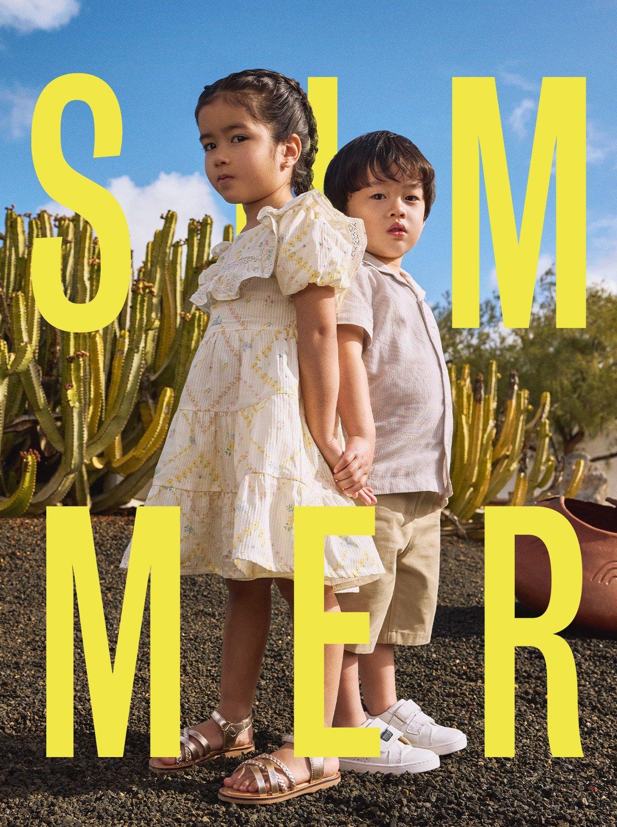 Girl in patterned summer dress and gold sandals, next to younger boy in light coloured short sleeved shirt, khaki shorts, and white velcro trainers