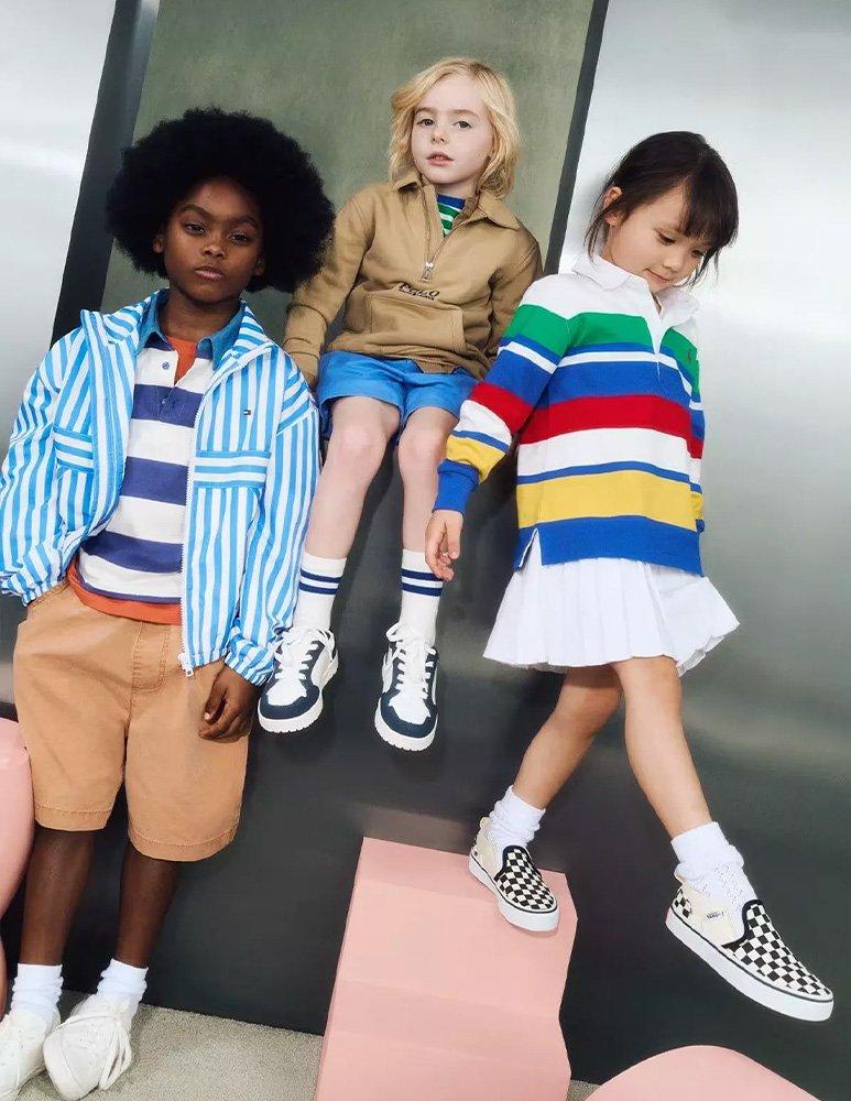 Boy standing wearing striped polo t-shirt with striped shirt over the top, khaki shorts and white trainers. Second younger boy sitting on a silver box wearing khaki jacket, blue shorts, striped skater socks and trainers. Young girl steppimg off a pink box wearing striped polo sweat shirt, white skirt, and checkerboard skater slip on sneakers