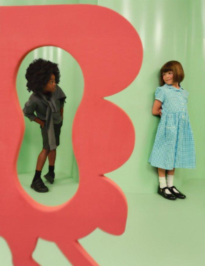 Two children playfully peek at each other around a large red cutout shape in a pastel green room.