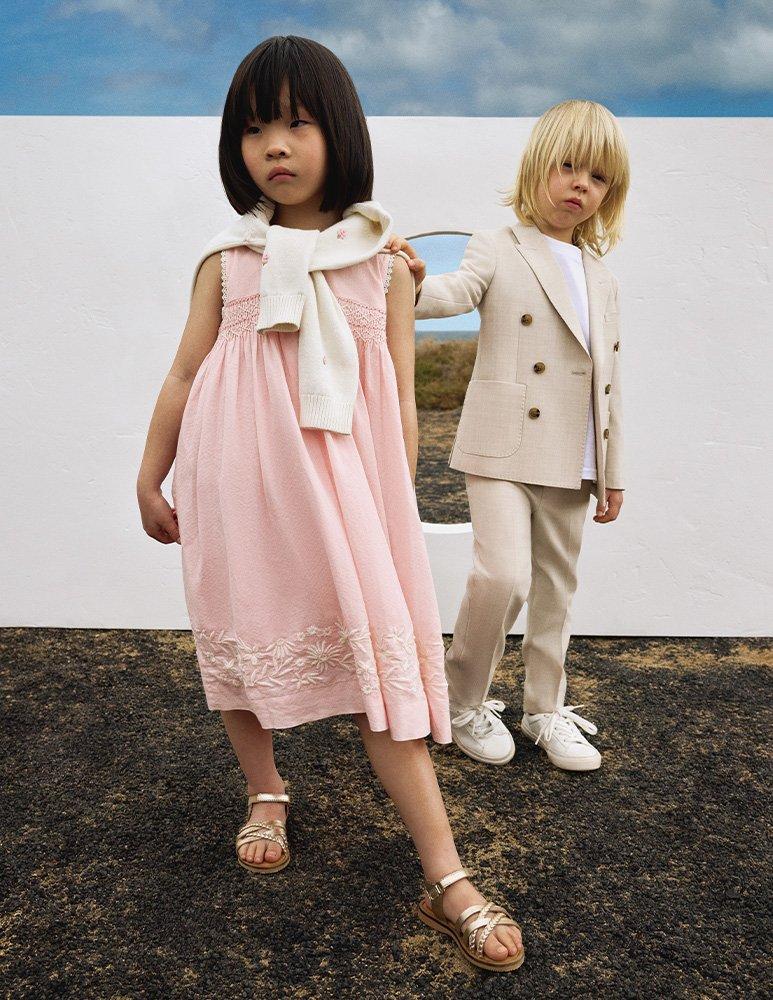 Girl in pink summer dress, gold sandles and jumper wrapped around shoulders, and boy in cream coloured suit jacket and trousers, and white trainers.