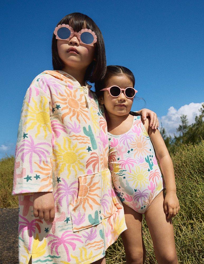 Girl in hooded towel palm tree patterned poncho and flower sunglasses, with younger girl in palm tree patterned swimming costume