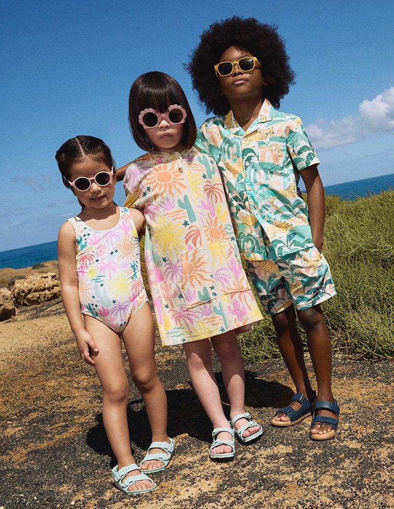 Three children wearing palm tree patterned swimming costume, palm tree patterned towel poncho, and palm tree matching set short sleeved shirt and shorts