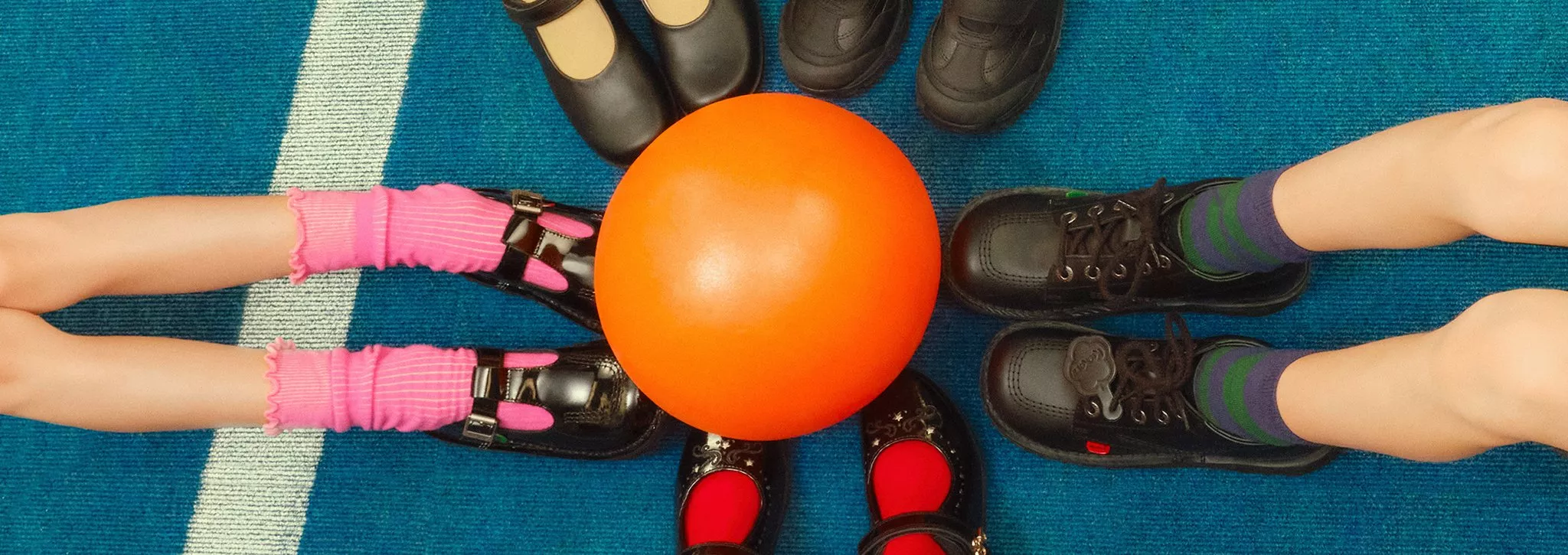 Children's feet in colourful socks and black shoes surround an orange ball on a blue carpet.