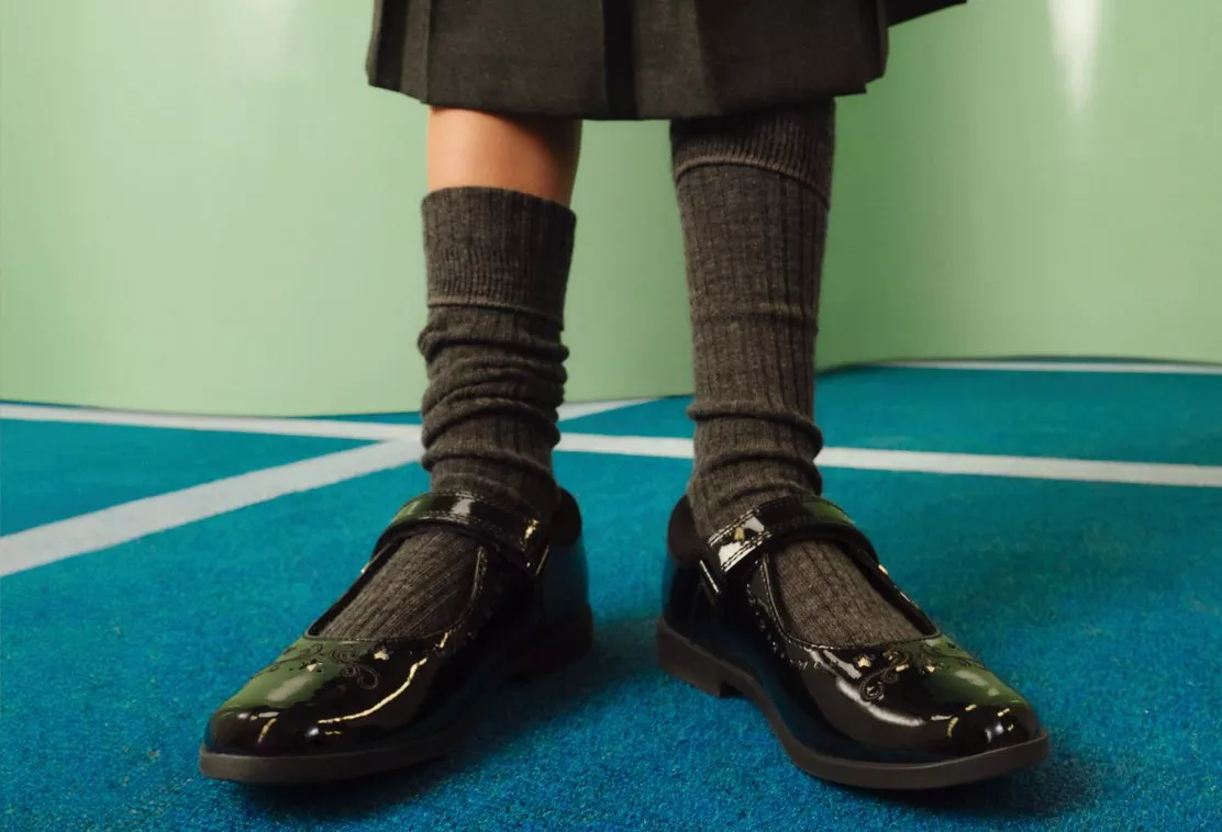 A close-up of a child wearing grey ribbed knee-high socks and shiny black patent leather shoes with embroidered details.