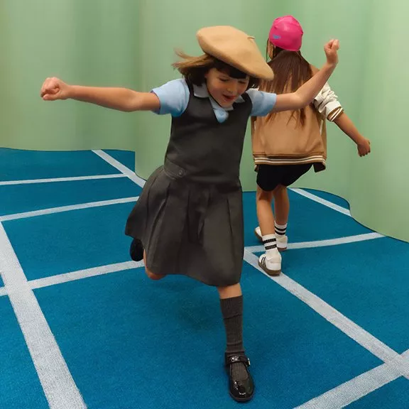Two children joyfully running indoors on a blue grid-patterned floor, one wearing a beret and school uniform.