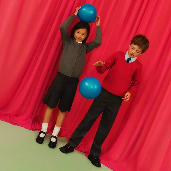 Two children in school uniforms play with blue balls against a vibrant pink curtain backdrop.