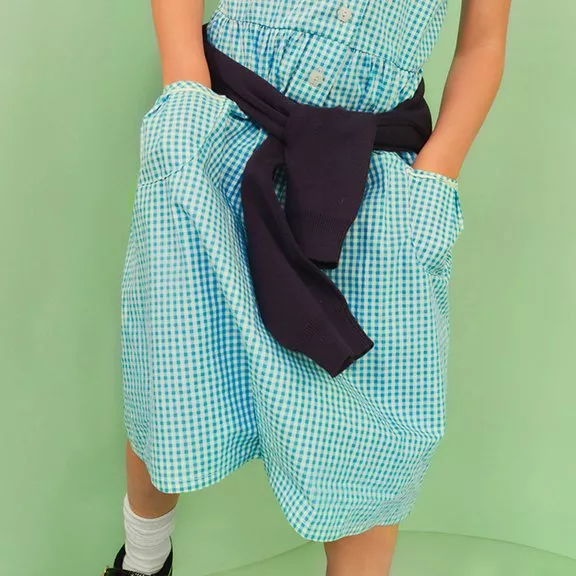 A child in a blue gingham dress with a navy jumper tied at the waist, standing against a green background.