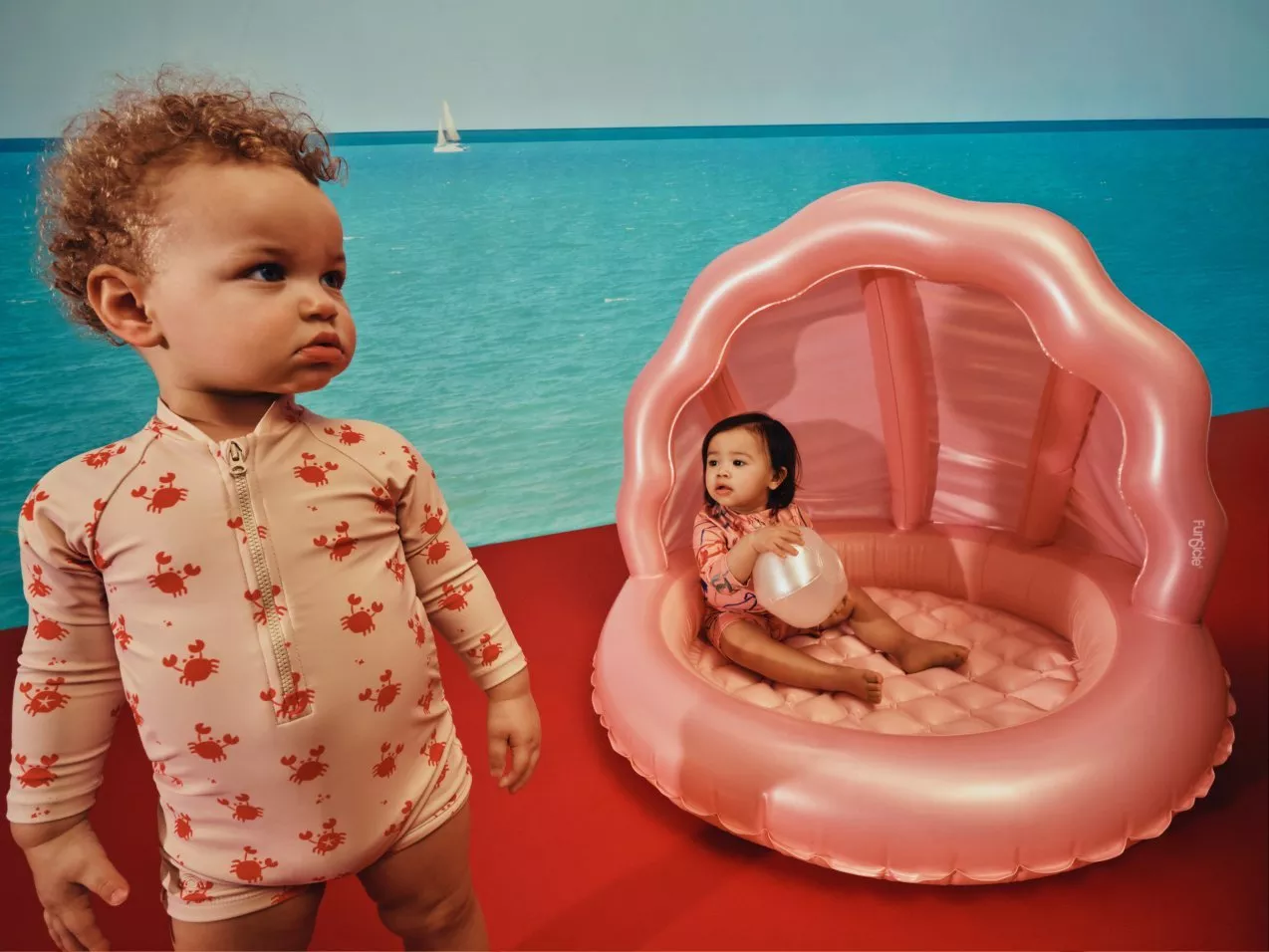 Two toddlers by the sea, one in a crab-print swimsuit, the other in a pink inflatable shell paddling pool.