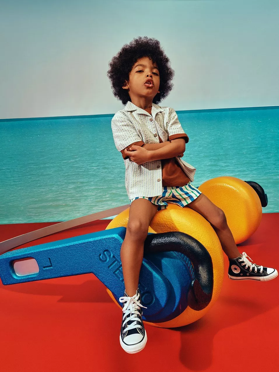 Boy in short sleeved shirt and swim shorts sitting on giant headphones, against a sea side backgraound