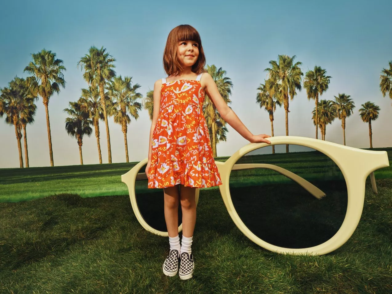 Girl in orange patterned dress posing beside oversized sunglasses on grass with palm trees backdrop