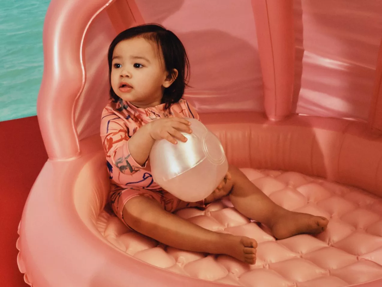 Toddler in pink swimwear holding a beach ball on a pink inflatable lounger beside a swimming pool