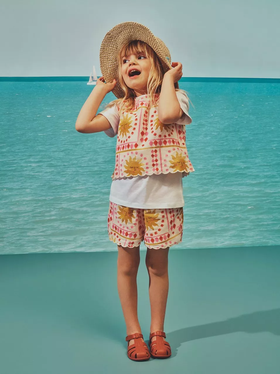 Child wearing a straw sunhat and patterned summer outfit, standing by turquoise sea background