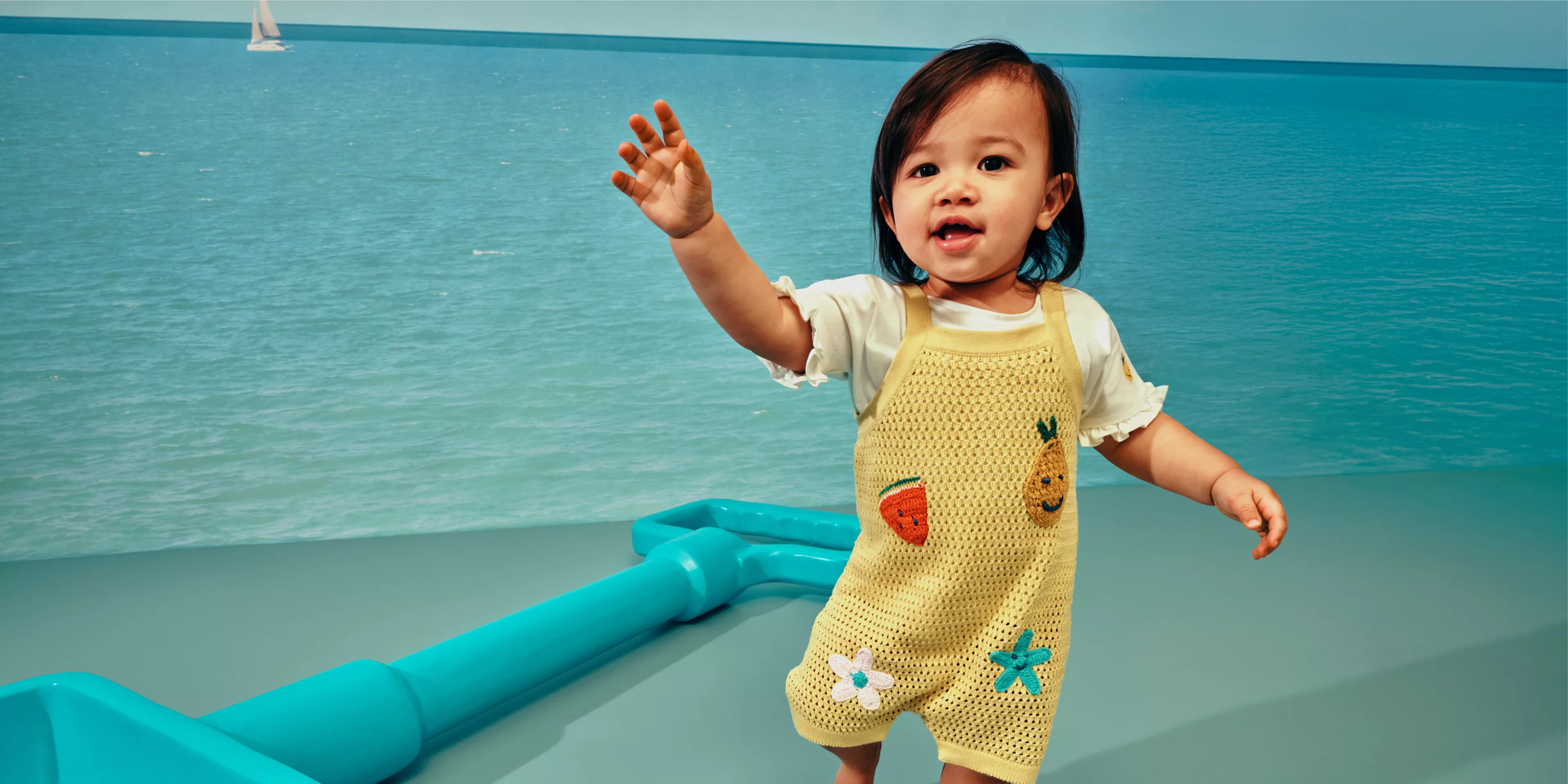 Toddler in yellow knitted dungarees with fruit motifs, against a sea side background