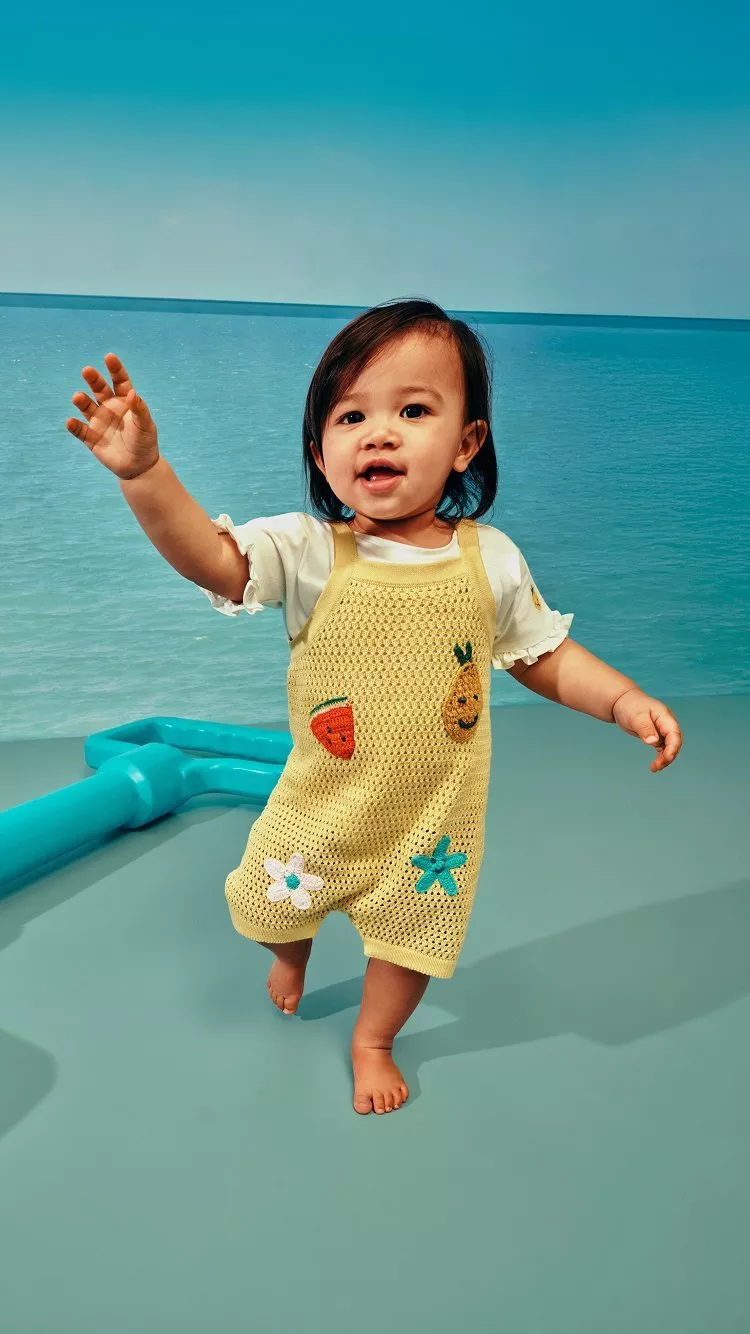 Toddler in yellow knitted dungarees with fruit motifs, against a sea side background
