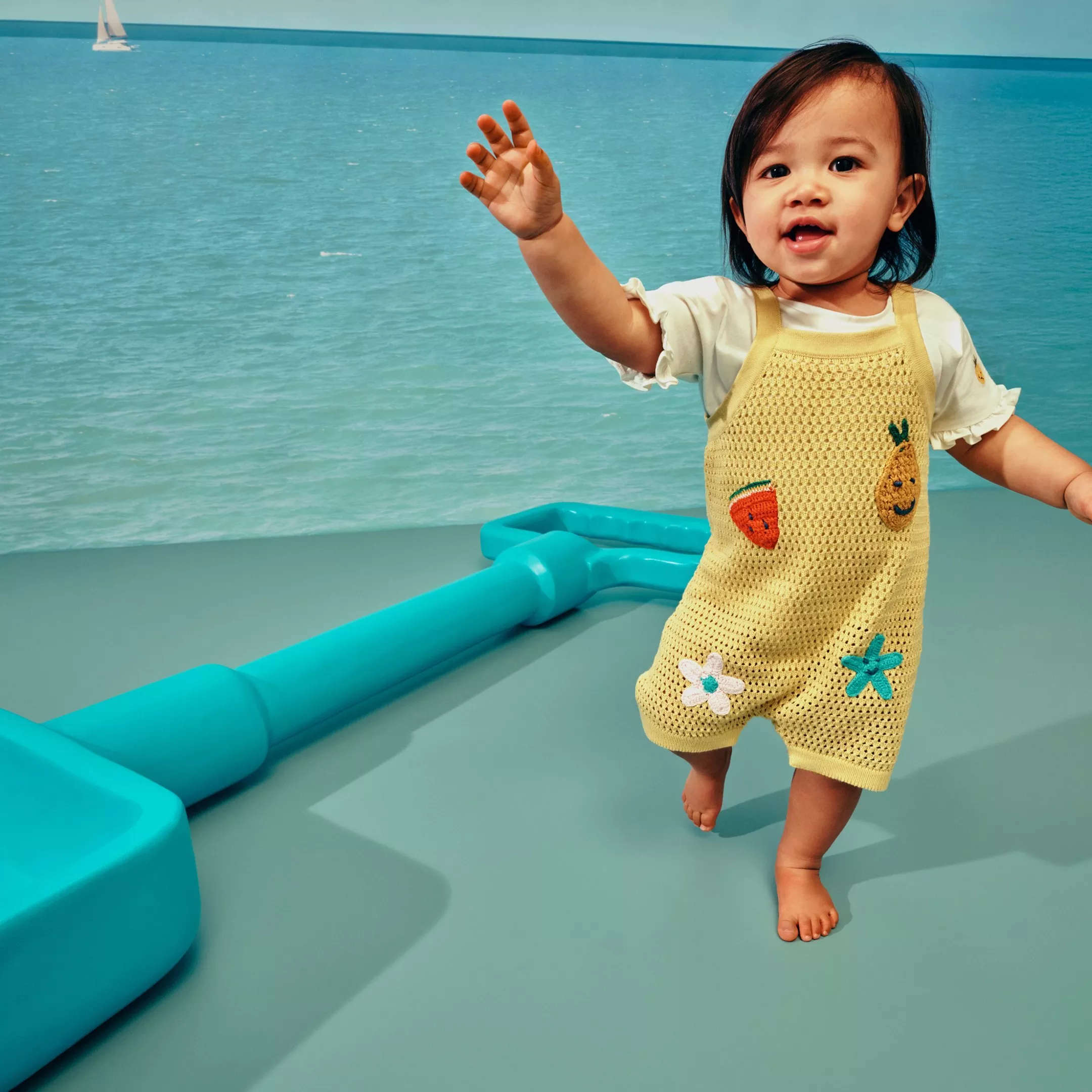 Toddler in yellow crochet romper with fruit patches, standing on deck by turquoise sea and distant sailboat.