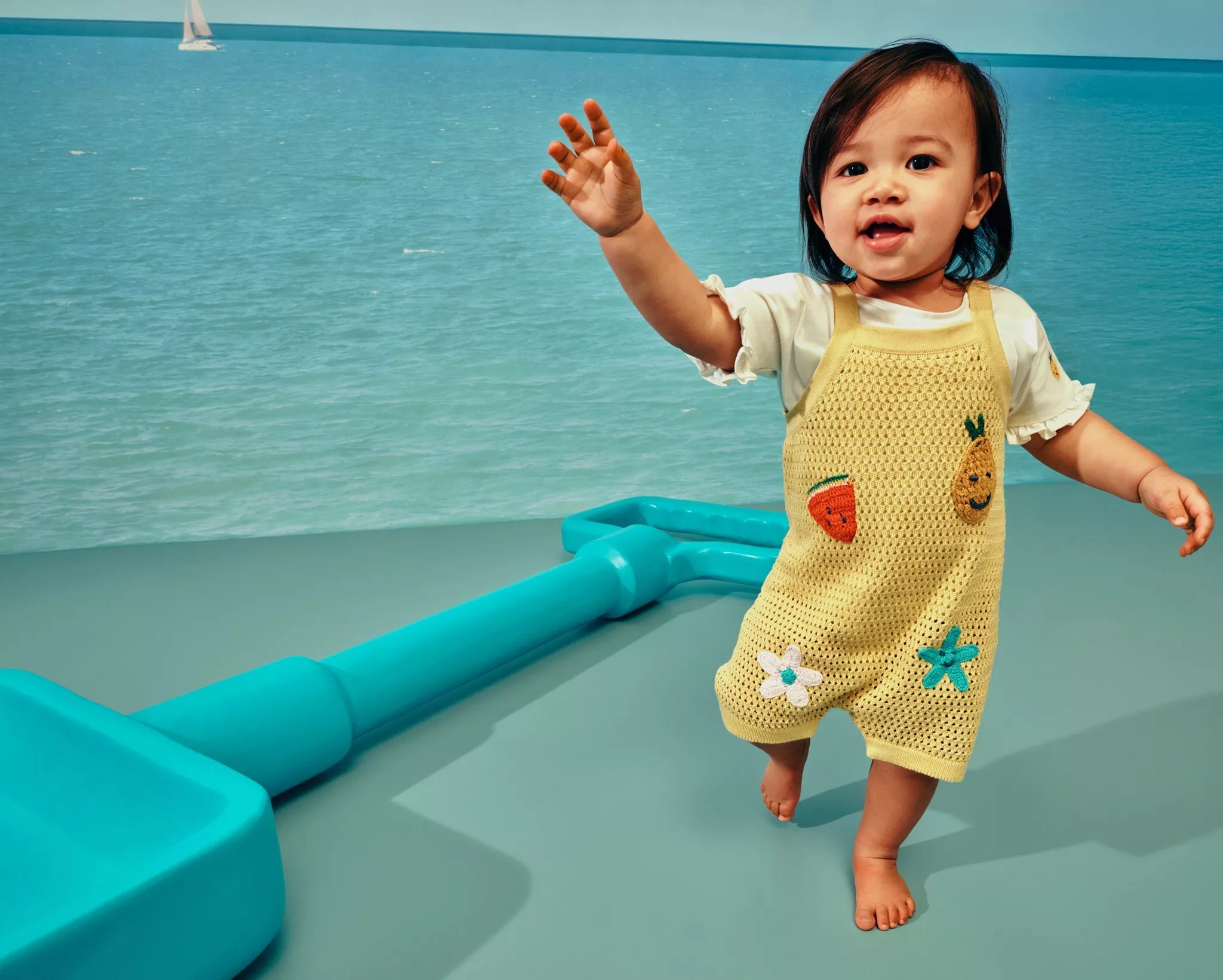Toddler in yellow knitted dungarees, against a seaside backdrop