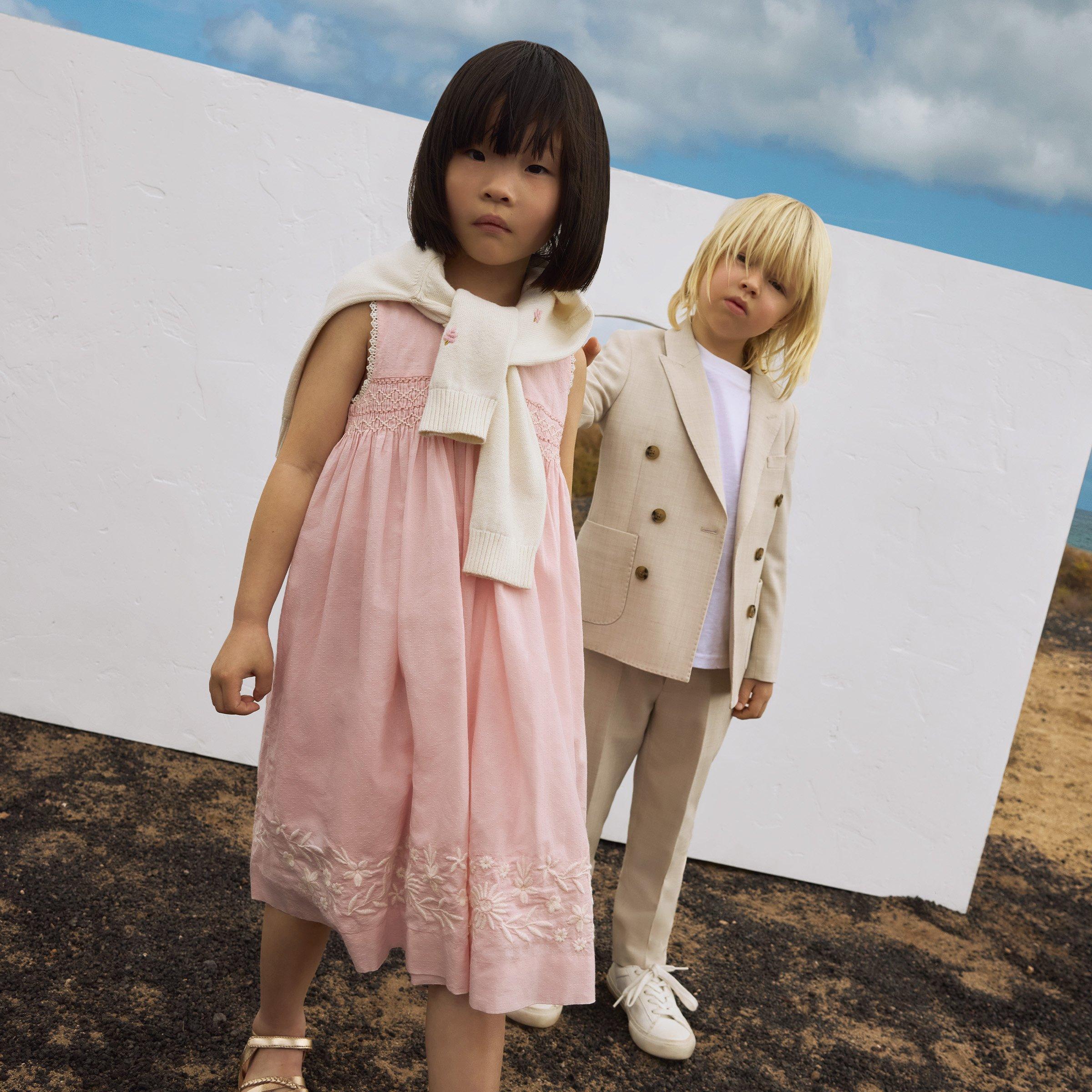 Girl in pink summer dress, gold sandles and jumper wrapped around shoulders, and boy in cream coloured suit jacket and trousers, and white trainers.