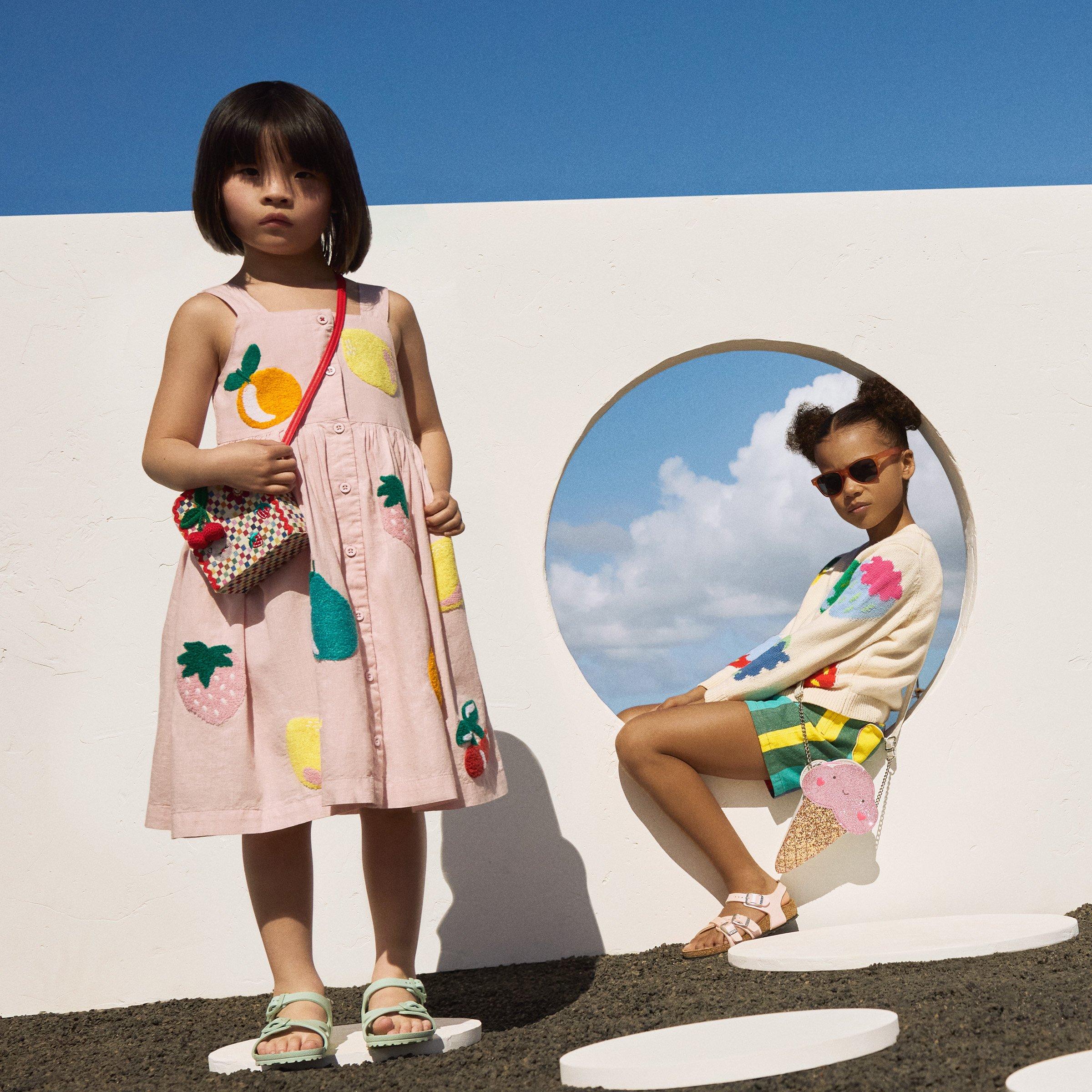 Two young girls in colorful, playful clothing with fruit and ice cream motifs, posing outdoors against a modern white wall with circular cutouts and a blue sky background.