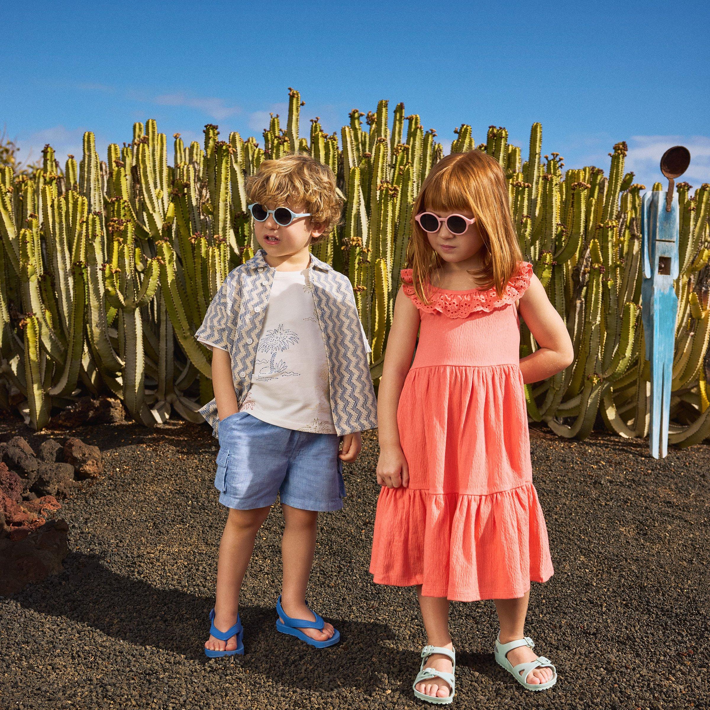 Two children standing outdoors in front of cacti, wearing sunglasses and summer clothes.