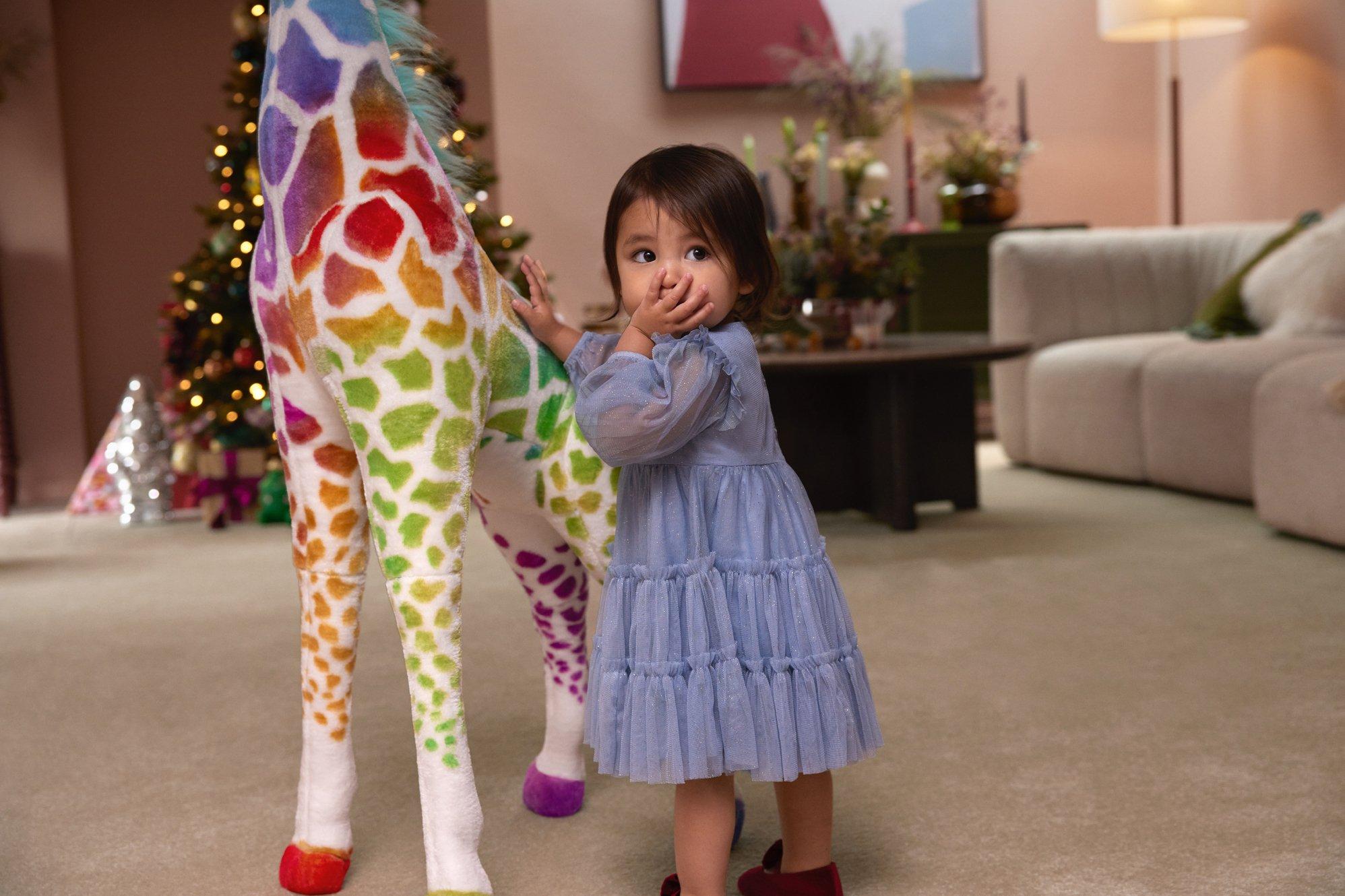 Image of a young girl next to a giant rainbow giraffe beside a Christmas treet