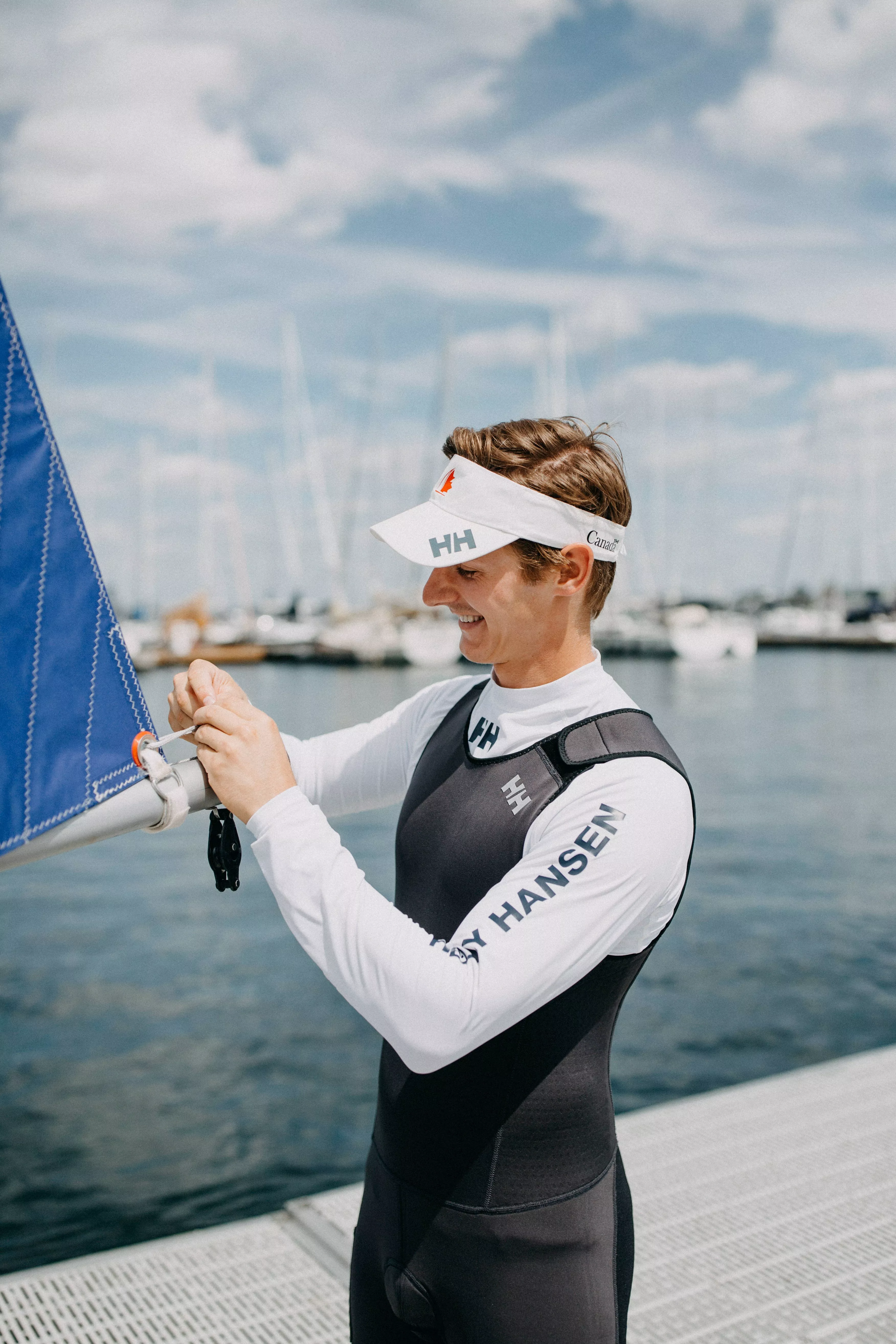 Sailor in wetsuit rigging a blue sail on a dinghy at a marina, preparing for sailing on calm water