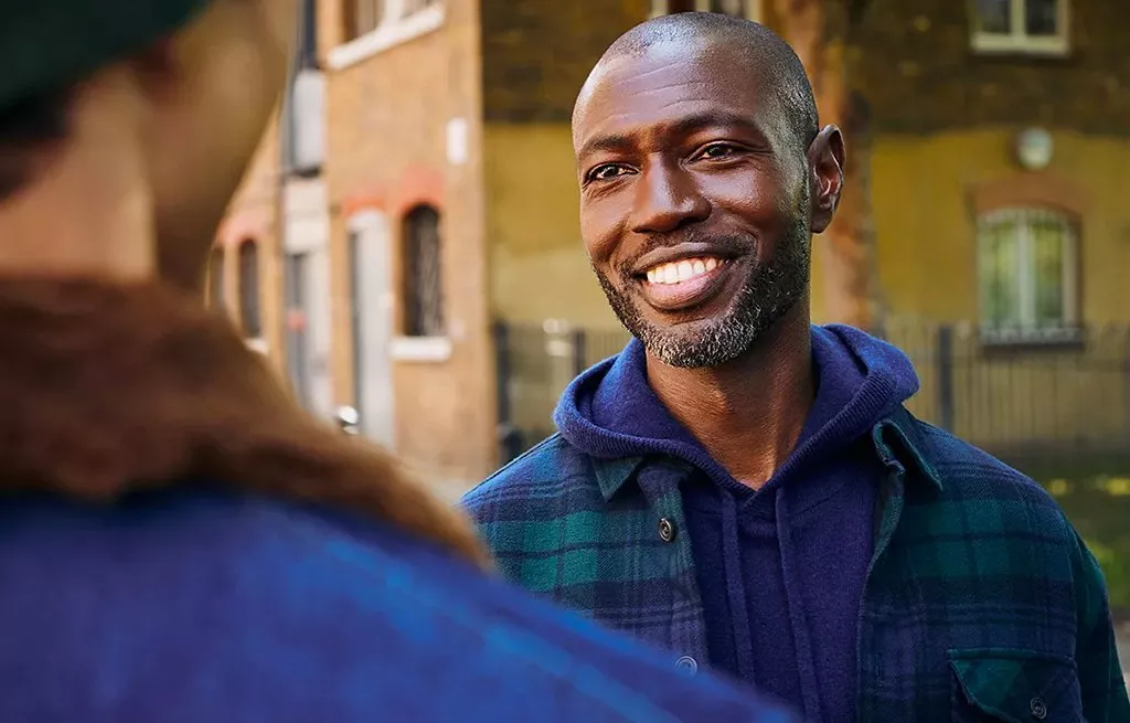 Smiling man in a hoodie and check shirt chatting outdoors on a city street, warm daylight and blurred background.