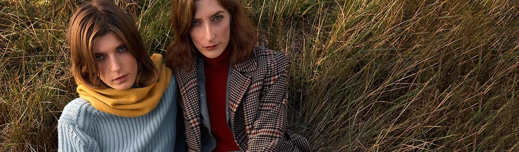 Two women in autumn knitwear and check coat sitting in tall grass, outdoor fashion portrait in warm light