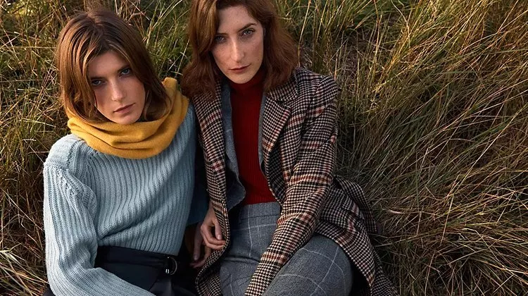 Two women in autumn knitwear and check coat sitting in tall grass, outdoor fashion portrait in warm light