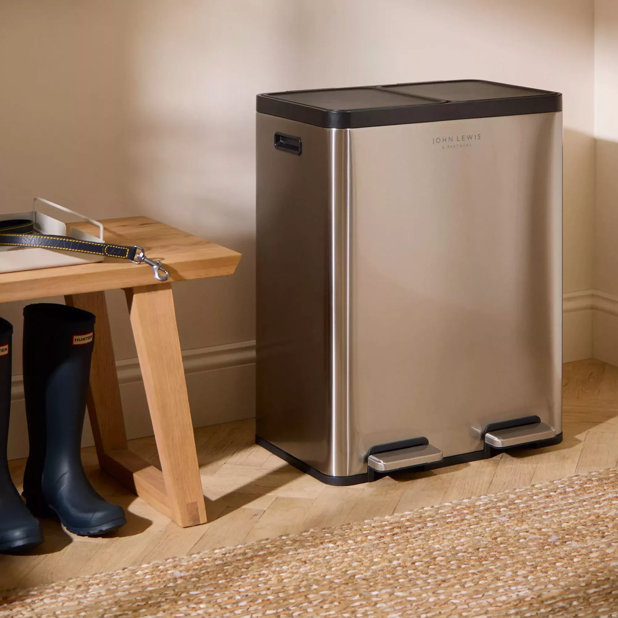Stainless steel dual-compartment pedal kitchen bin beside wooden bench in hallway, ideal for recycling and waste