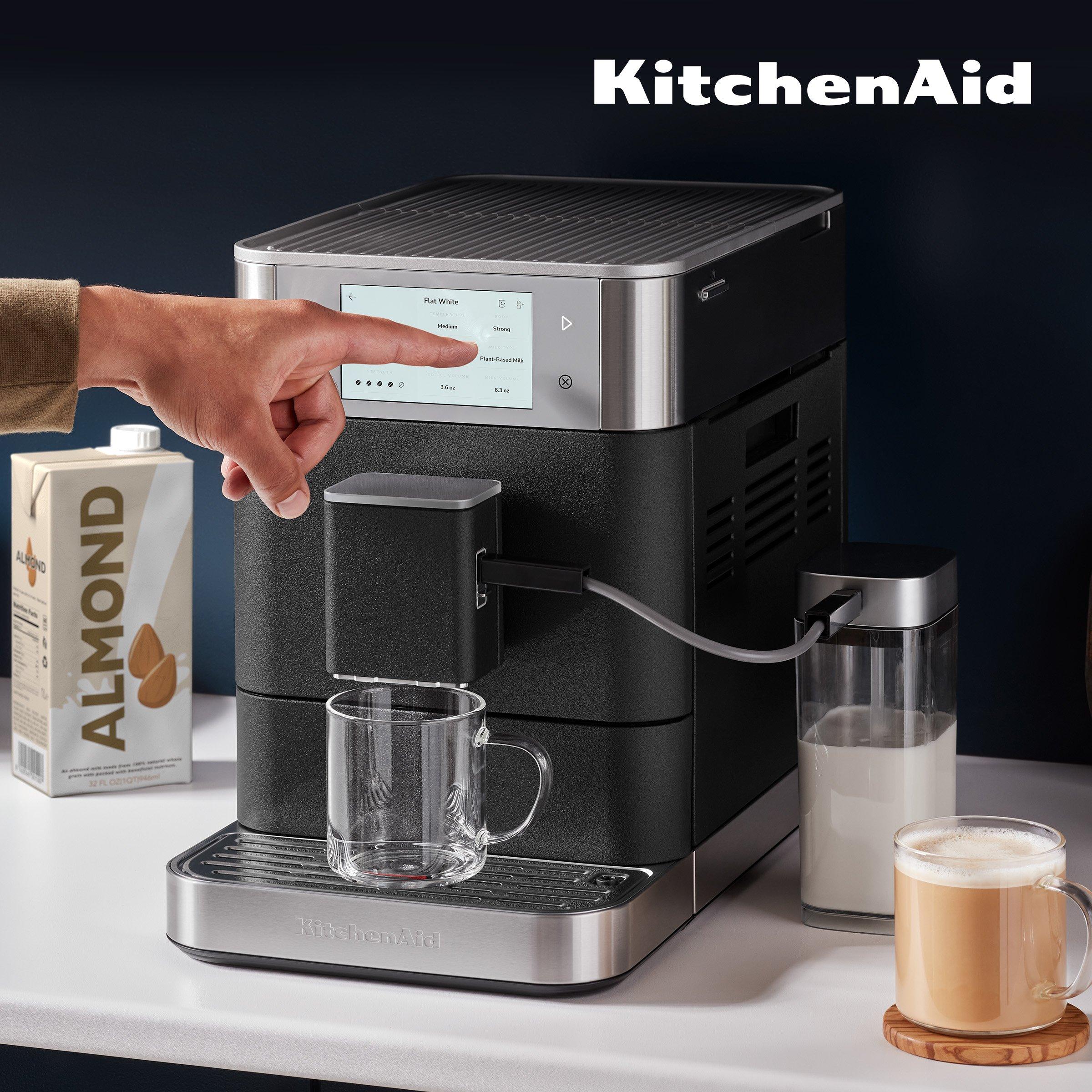 A KitchenAid automatic espresso machine with Glass Mug, Milk Container, Carton of Almond Milk. A hand is actively using the touchscreen interface.