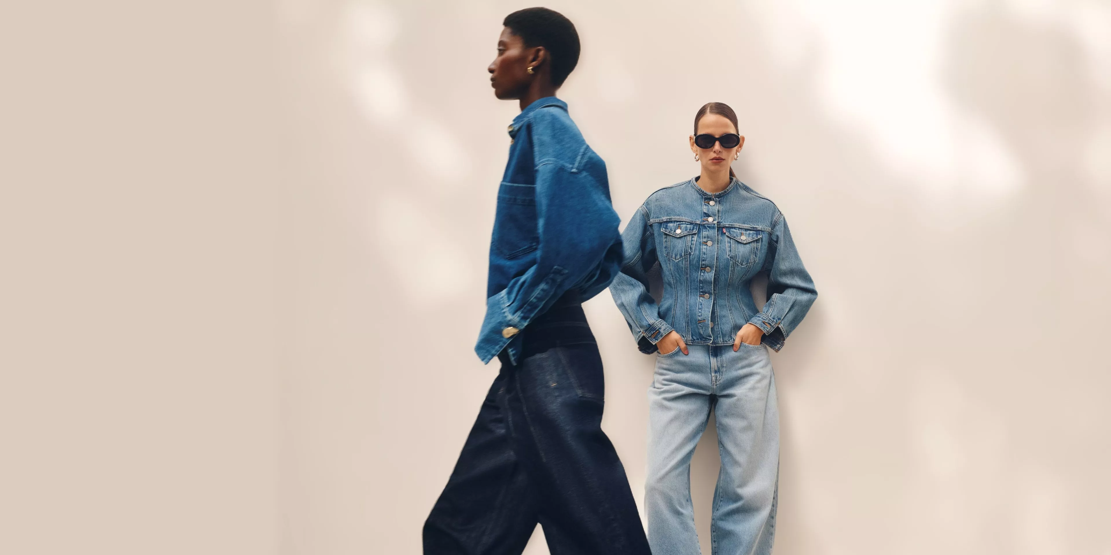 Two models in denim outfits; one walks by, the other leans against a wall wearing sunglasses, exuding a cool vibe.