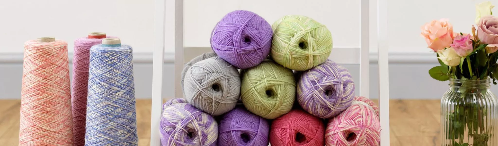 Colourful knitting yarn balls and cones on a craft shelf beside a vase of roses on a wooden table