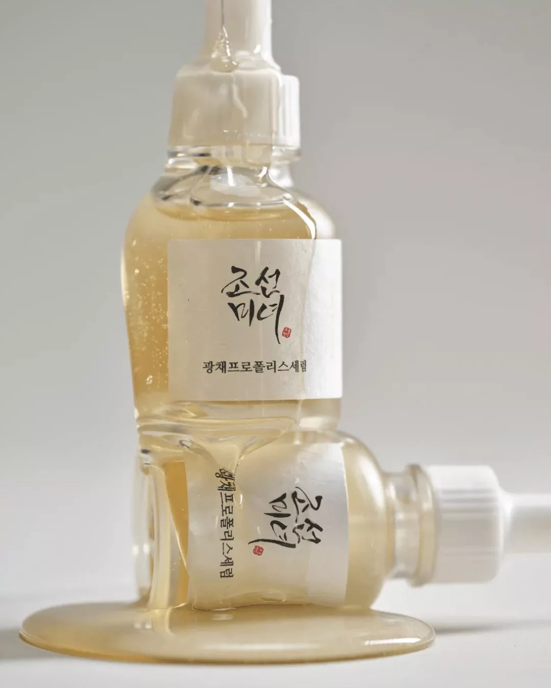 Close-up of Korean propolis serum dropper bottles with golden gel spill, minimalist skincare product shot