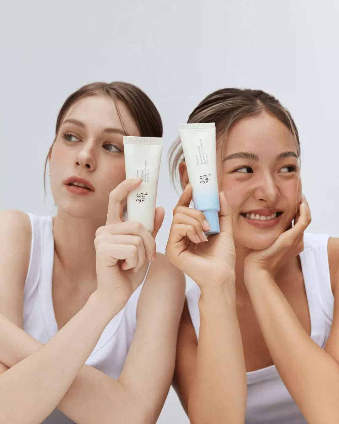 Two women holding skincare cream tubes, clean beauty product photo for moisturiser and face care routine.