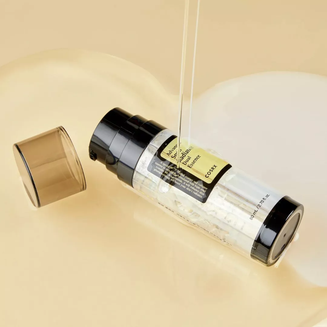 COSRX snail mucin radiance essence bottle with cap off, lying in creamy liquid skincare backdrop
