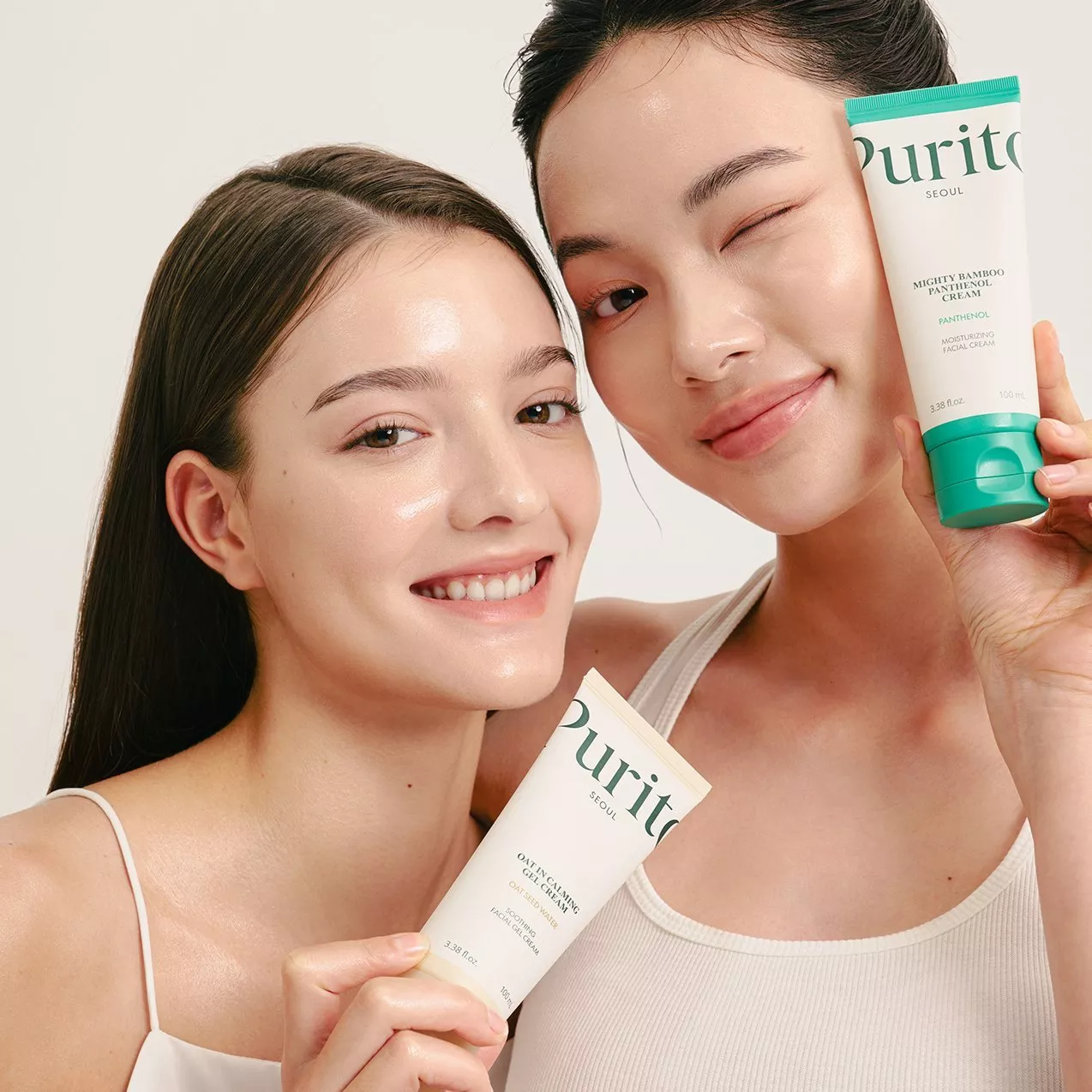 Two women with glowing skin holding Purito Seoul skincare tubes, smiling and winking in a studio portrait.