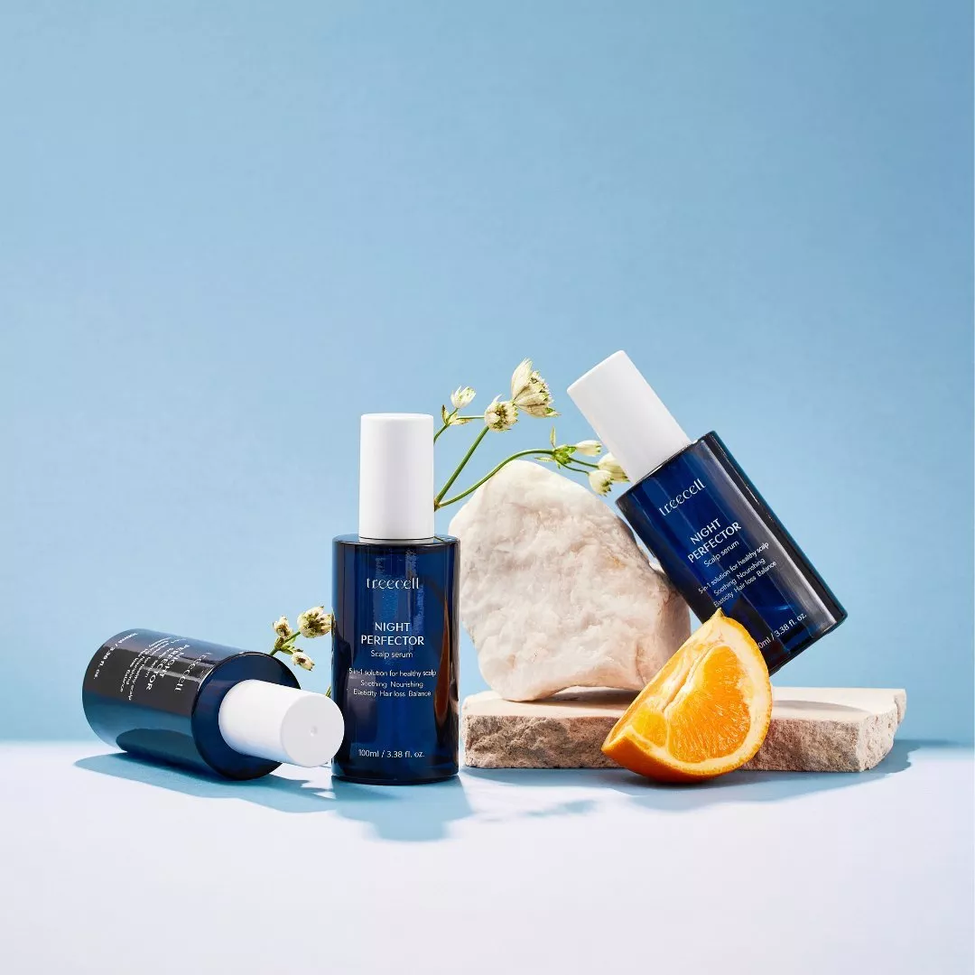 Three blue scalp serum bottles with white caps, styled with orange slice, flowers and stone on blue backdrop