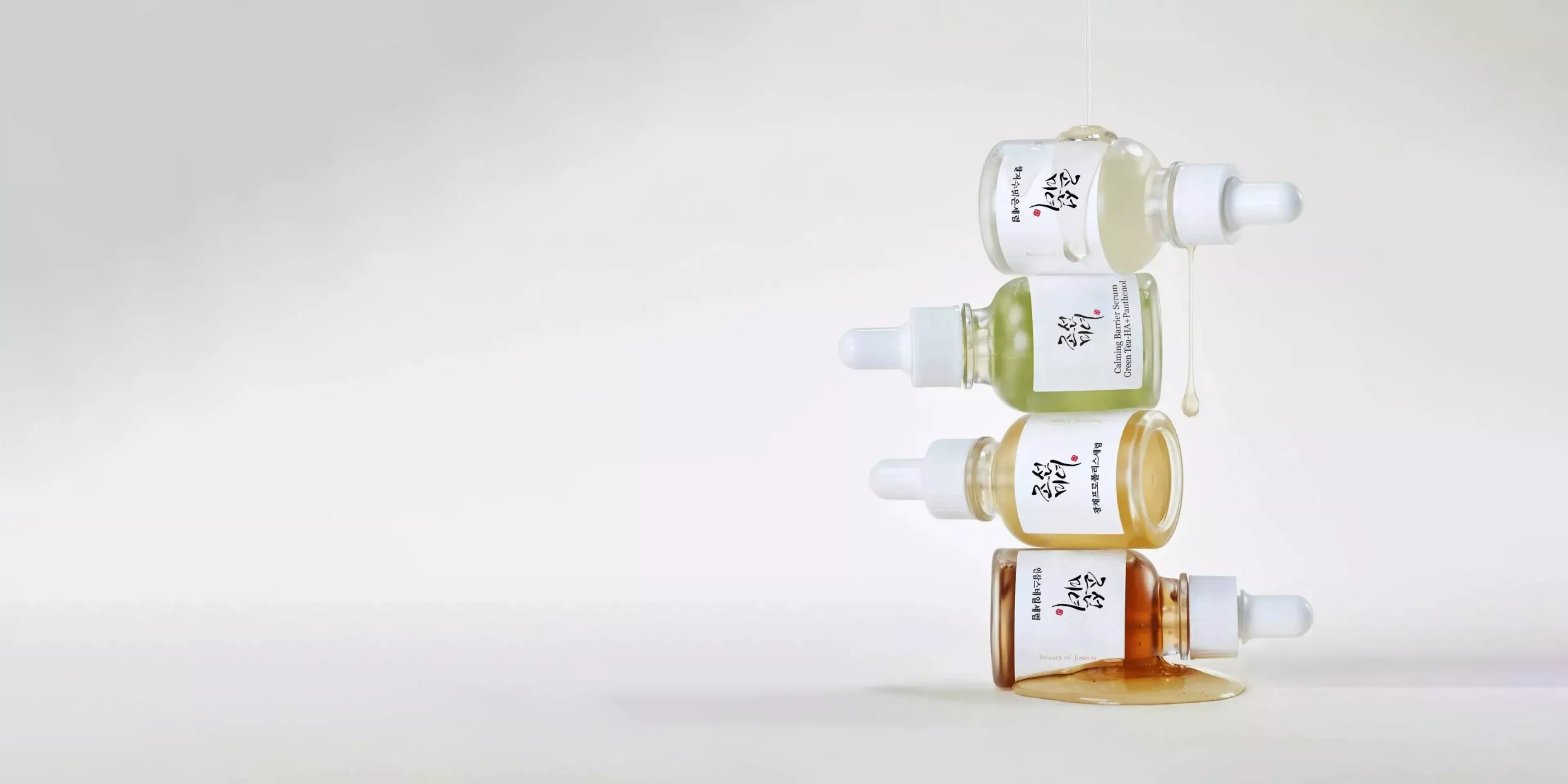 Stacked Korean skincare serum dropper bottles with spilled oil, minimalist product photography on white background