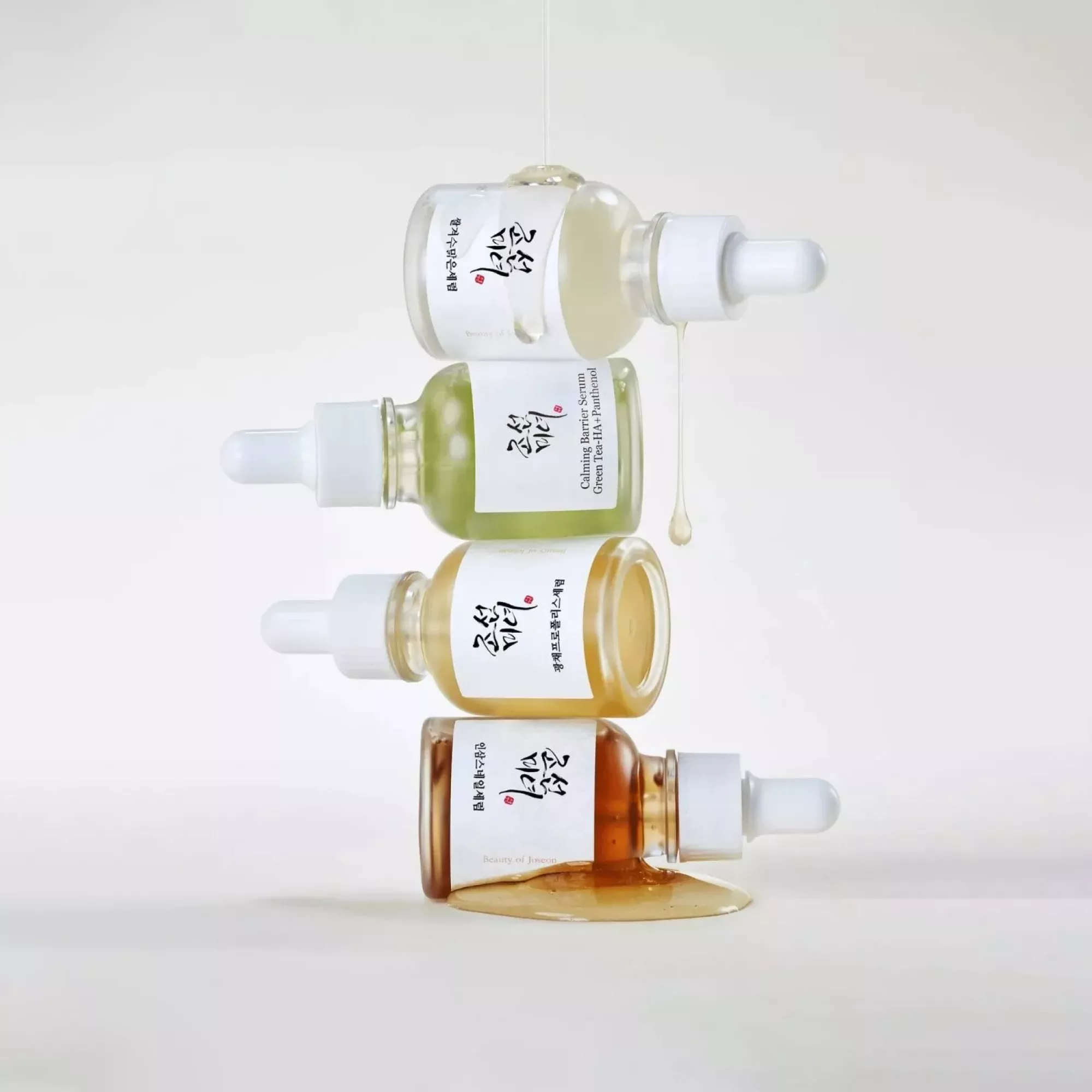 Stacked Beauty of Joseon serum dropper bottles with honey and green tea skincare oils dripping on white background