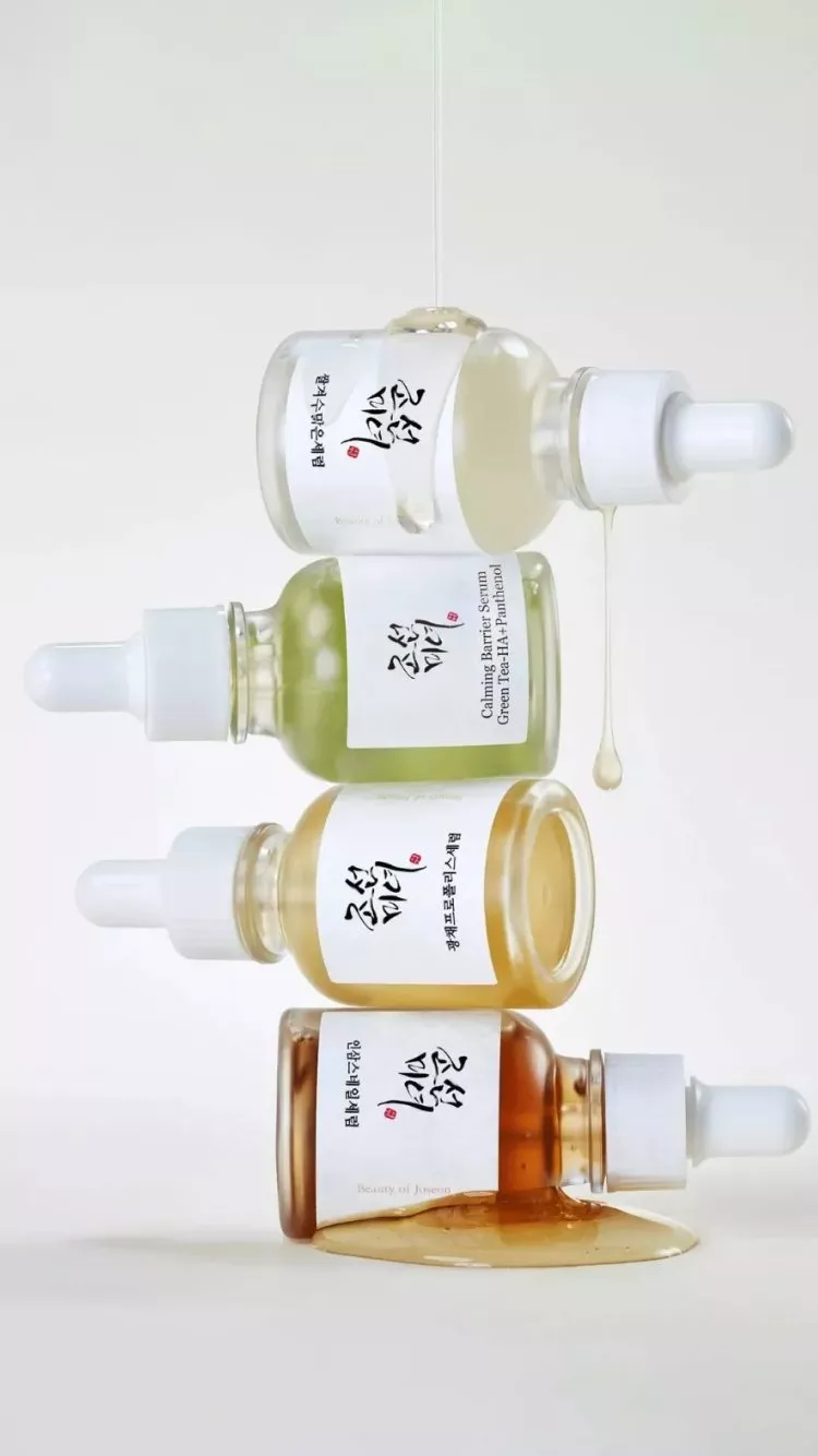 Stacked glass dropper bottles of Korean skincare serums, with liquid dripping and spilling on a white backdrop