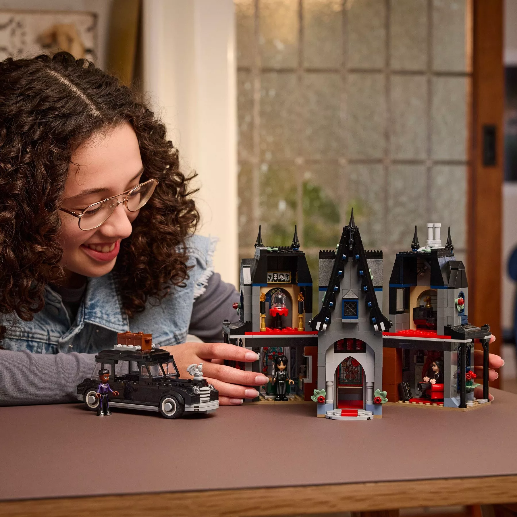 a girl with curly hair and glasses smiles while playing with a LEGO Wednesday Addams set featuring a black car and a detailed Victorian-style mansion