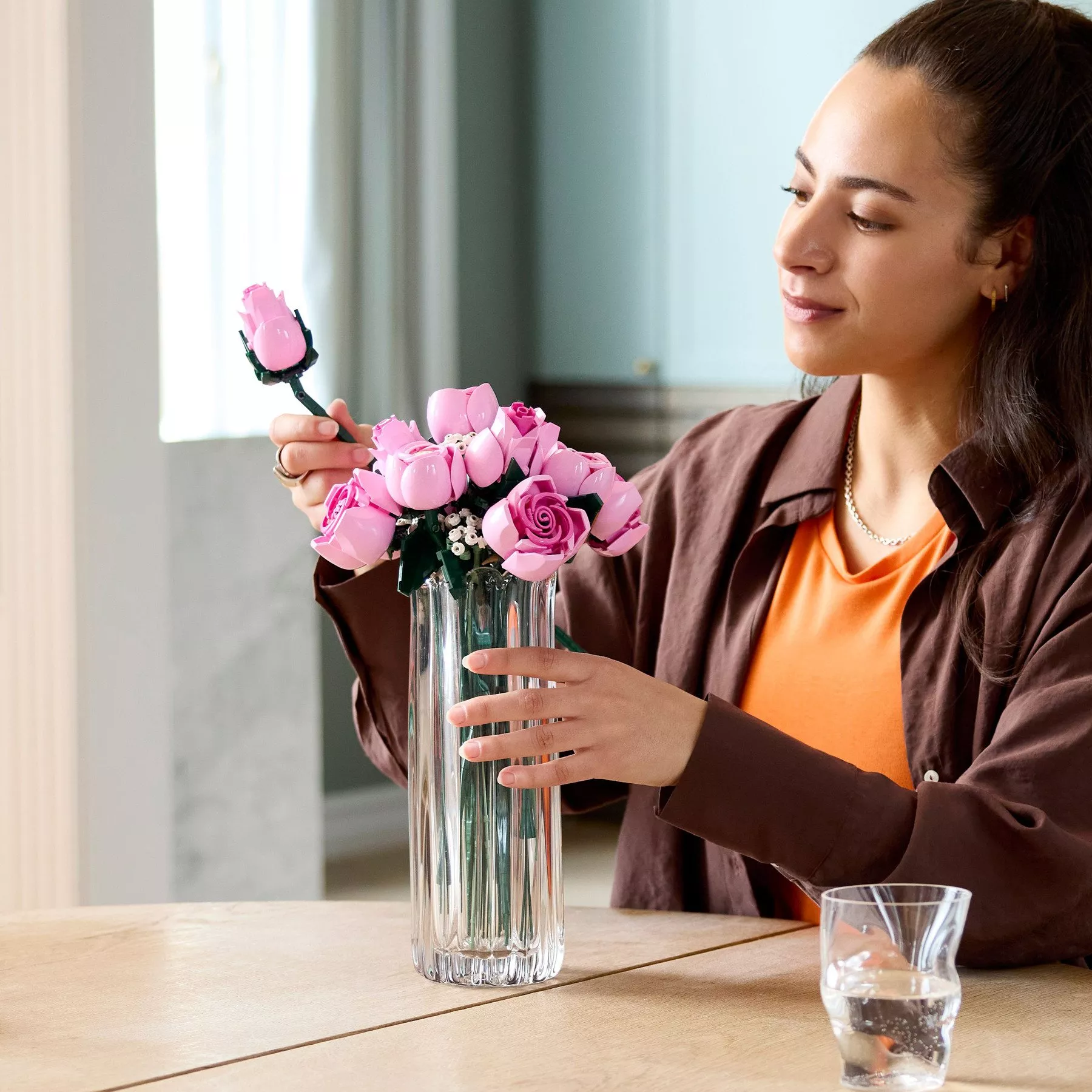 a woman with long dark hair smiles while arranging a bouquet of pink LEGO roses into a tall, clear fluted glass vase on a wooden table